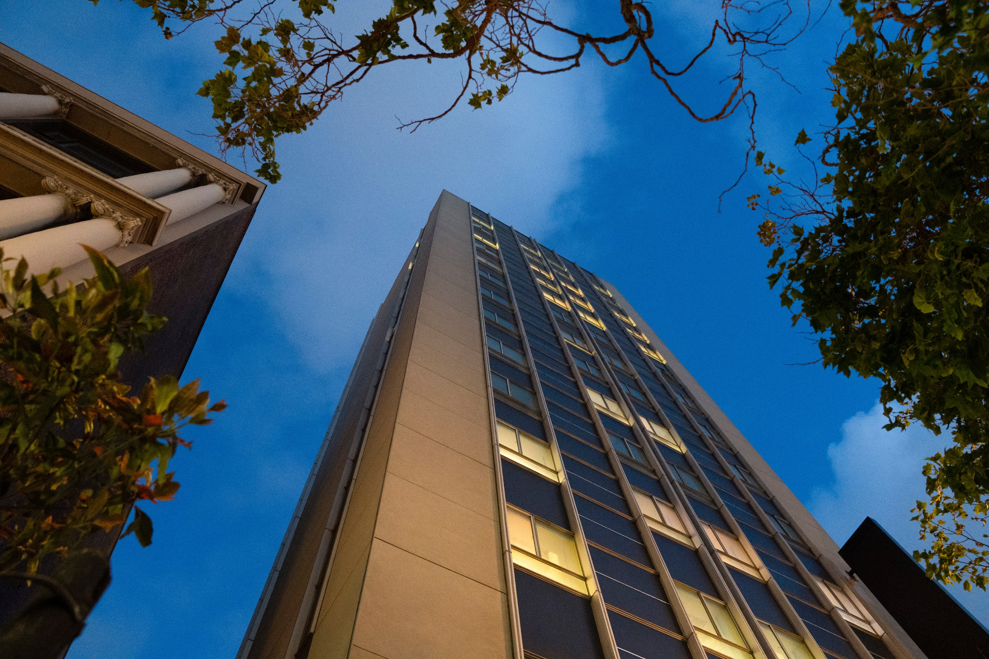 A tall, modern building is seen from below against a twilight sky. Trees frame the image, and the building has many lit windows, giving a sense of urban vibrancy.