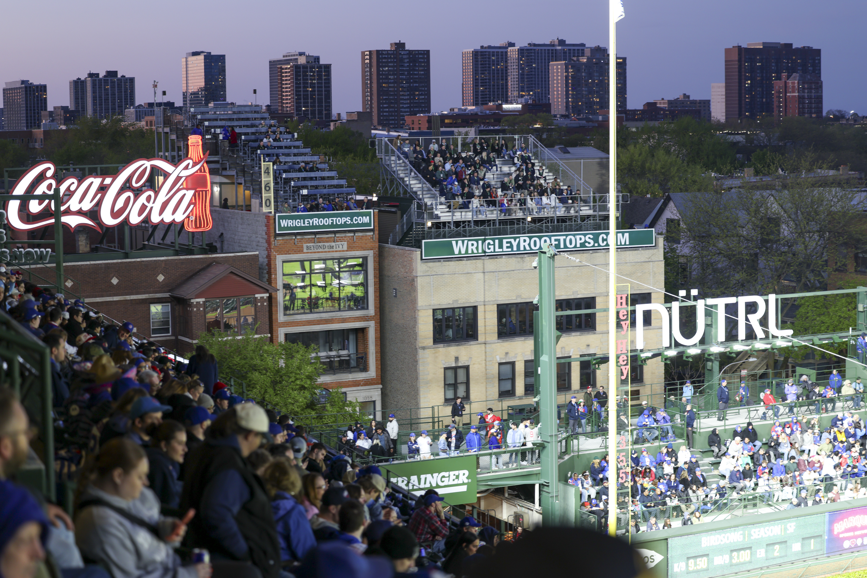 The image shows a crowded baseball stadium with rooftop seating in the background. There are bright signs, including a Coca-Cola and NÜTRL ad, and city buildings in the distance.