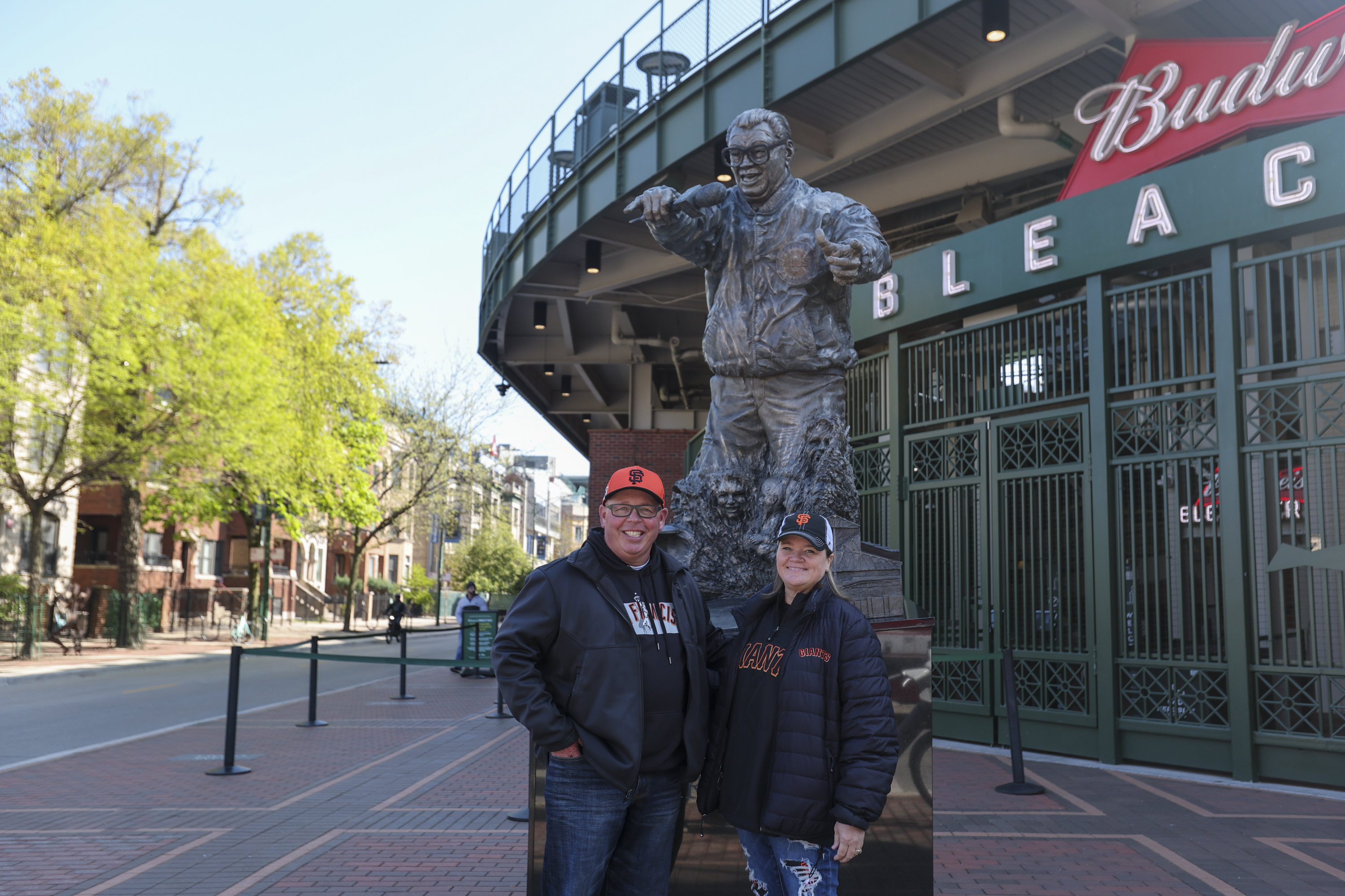 A couple poses in front of a bronze statue of a man holding a microphone outside a stadium entrance. They are wearing matching baseball team attire.