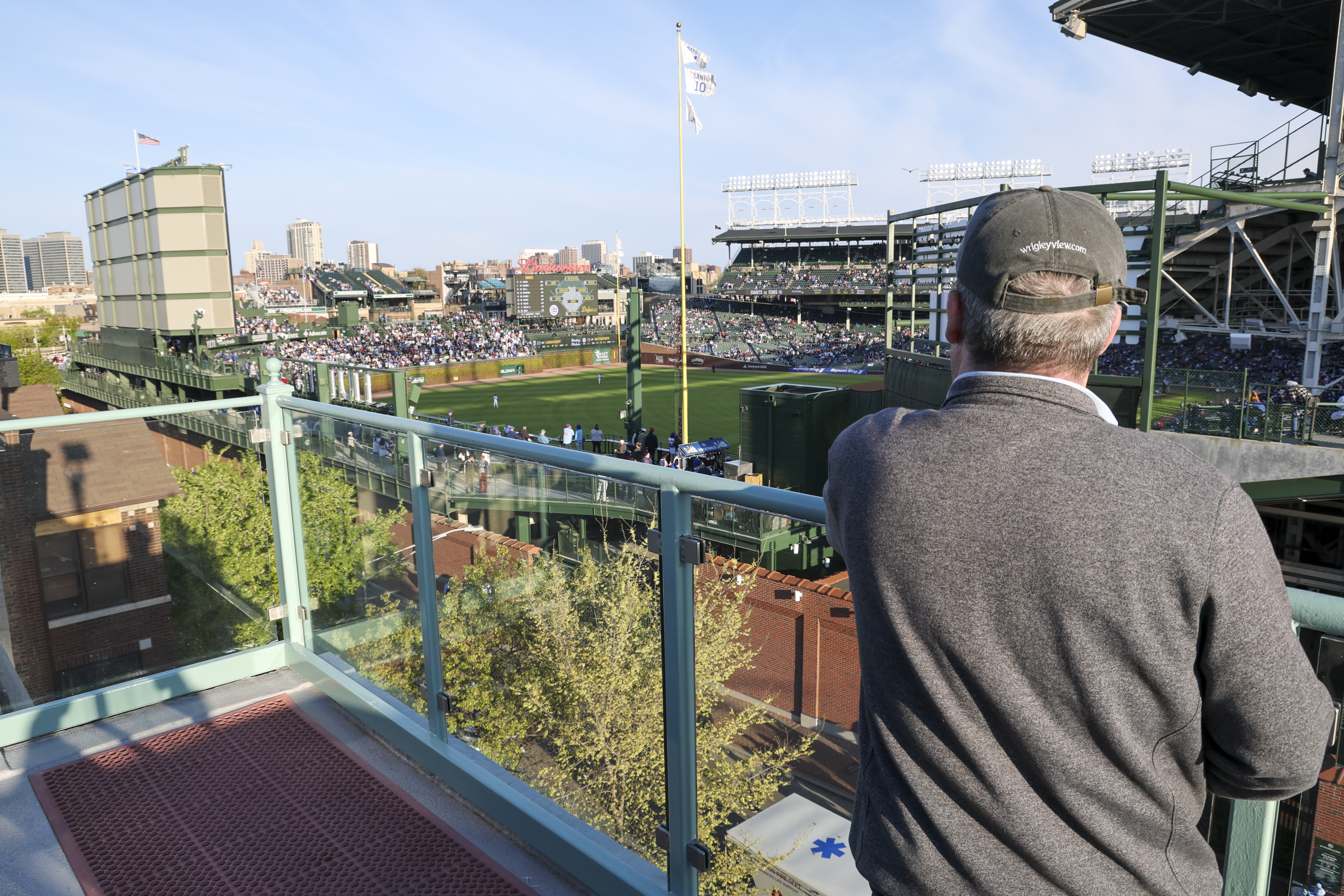 A man in a cap and sweater leans on a railing, overlooking a busy baseball stadium with people in the stands and a city skyline in the background.