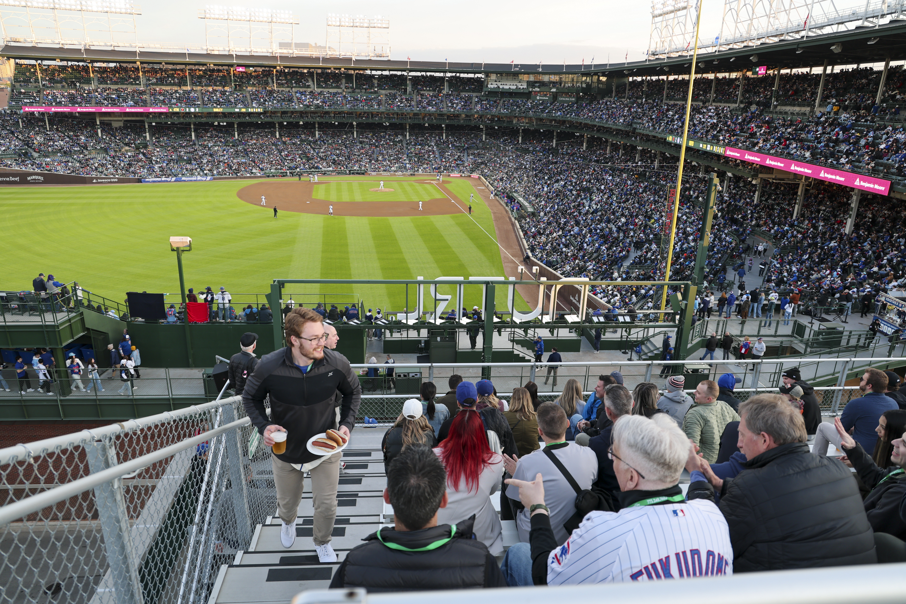 A crowded baseball stadium with a game in progress. Spectators fill the stands, while a man walks upstairs holding food and drinks. The field is brightly lit.