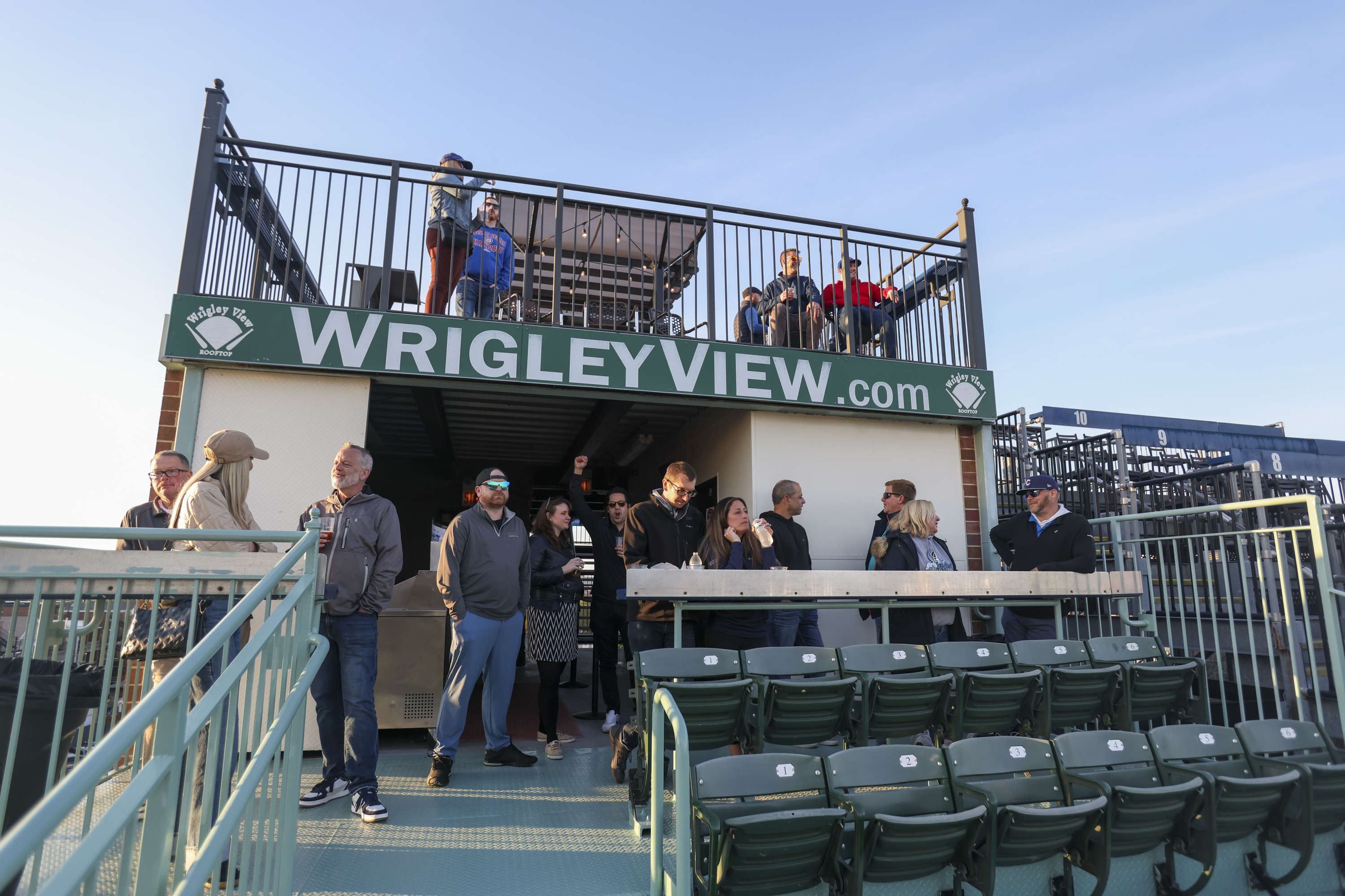 A group of people stand and socialize on a rooftop with a sign saying &quot;WrigleyView.com.&quot; There are green bleacher seats and metal railings visible.