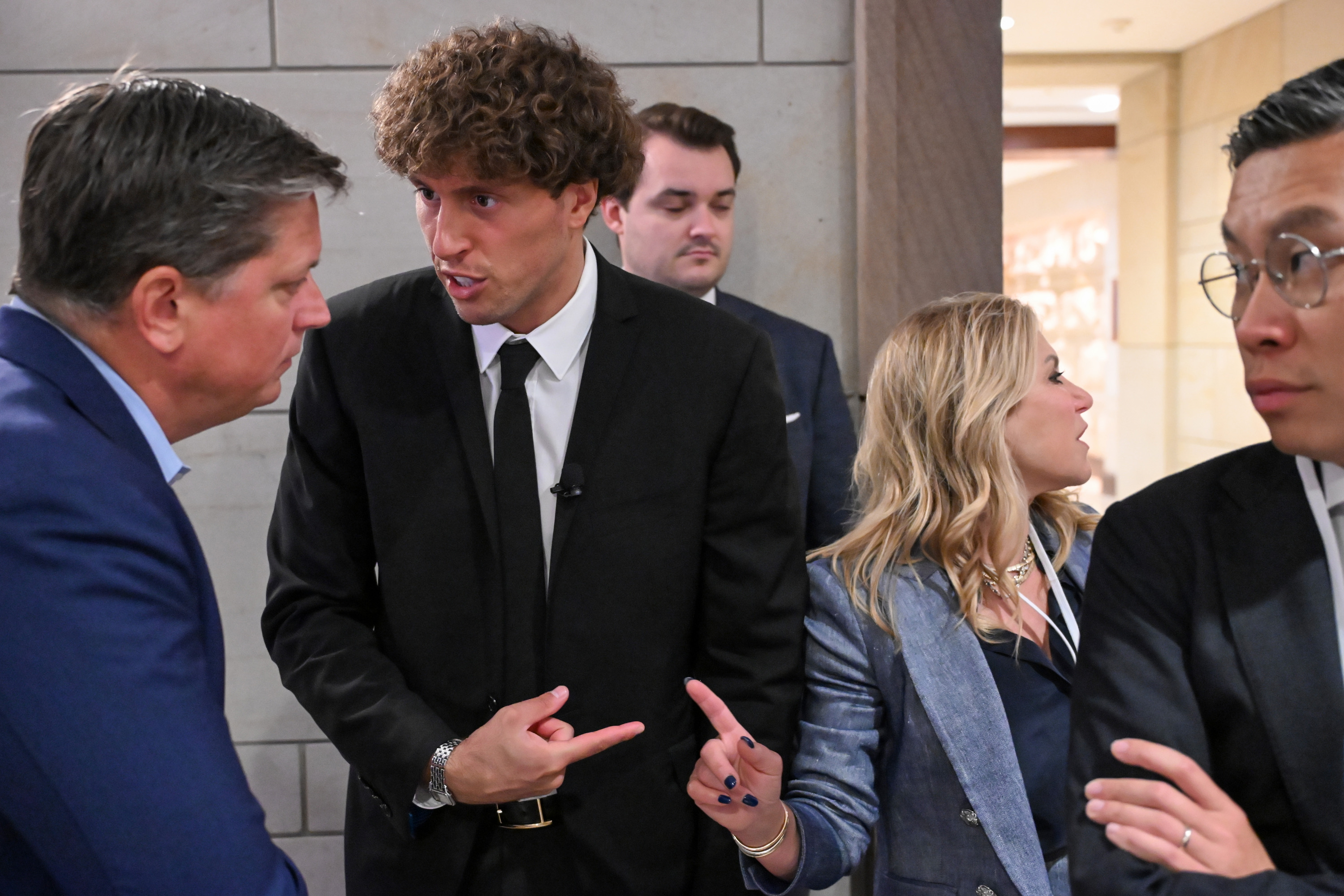 A group of people in suits are engaged in a conversation. One person, with curly hair, gestures while talking to another man in a blue suit. Other individuals look on.