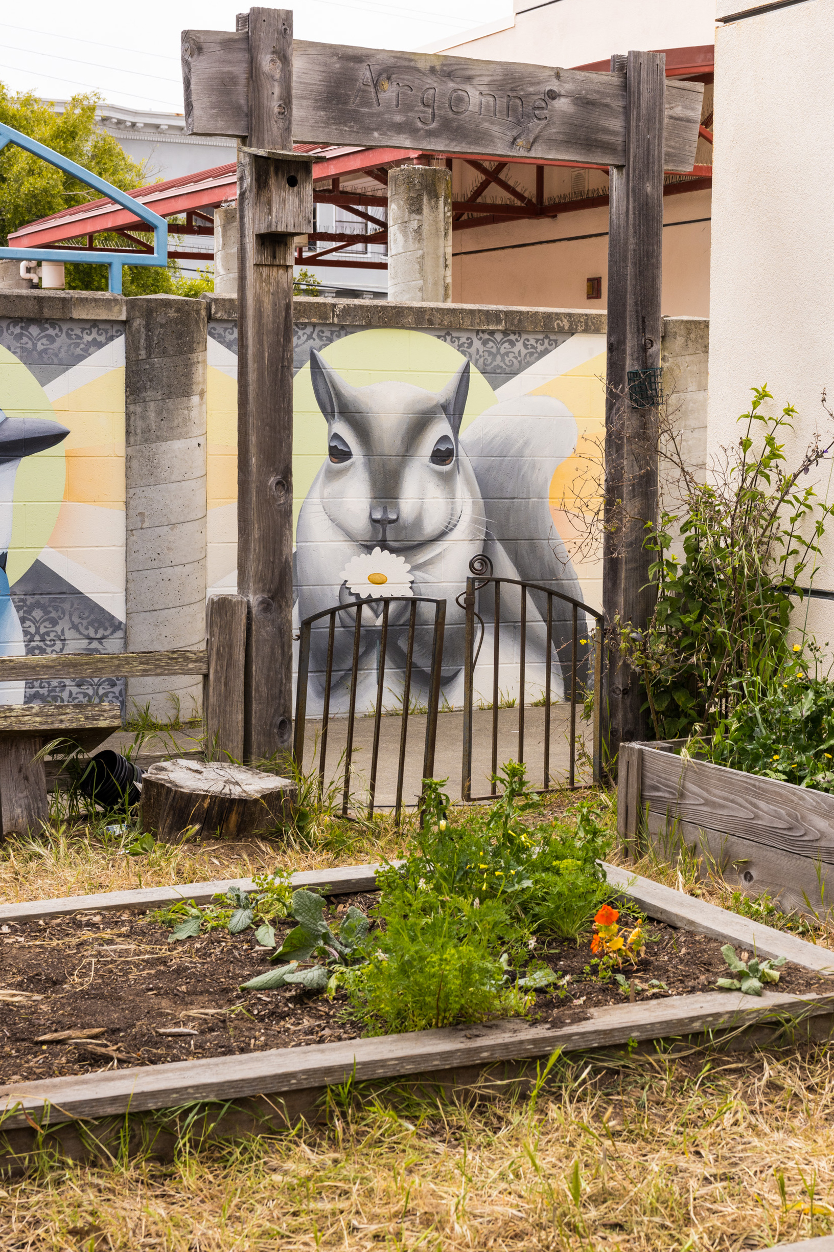 A garden scene shows a mural of a squirrel holding a flower on a wall, framed by wooden posts labeled &quot;Argonne,&quot; with a small garden bed in the foreground.