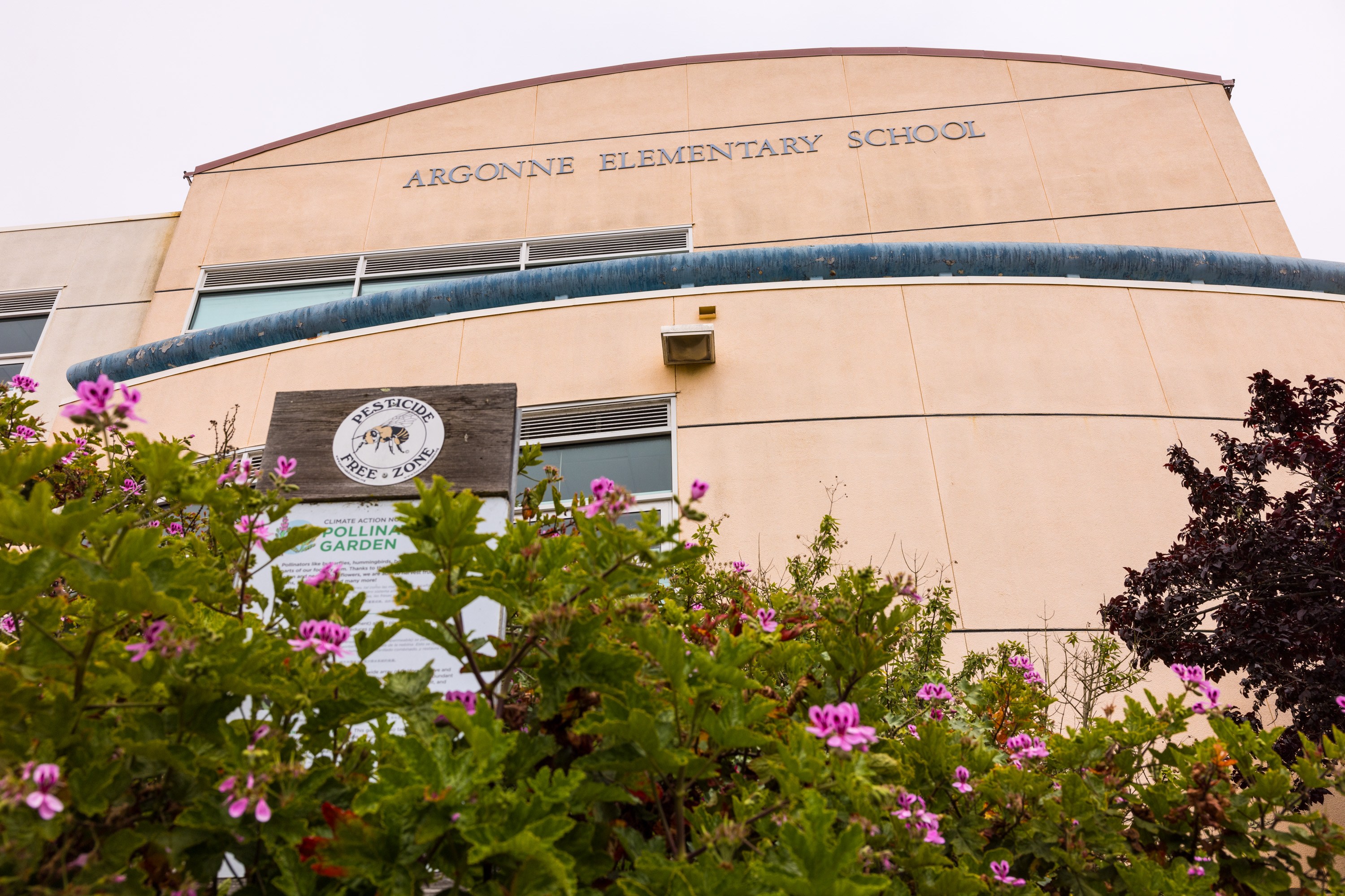 The image shows Argonne Elementary School's curved facade, with "Pesticide Free Zone" and garden signs, framed by blooming pink flowers and greenery.