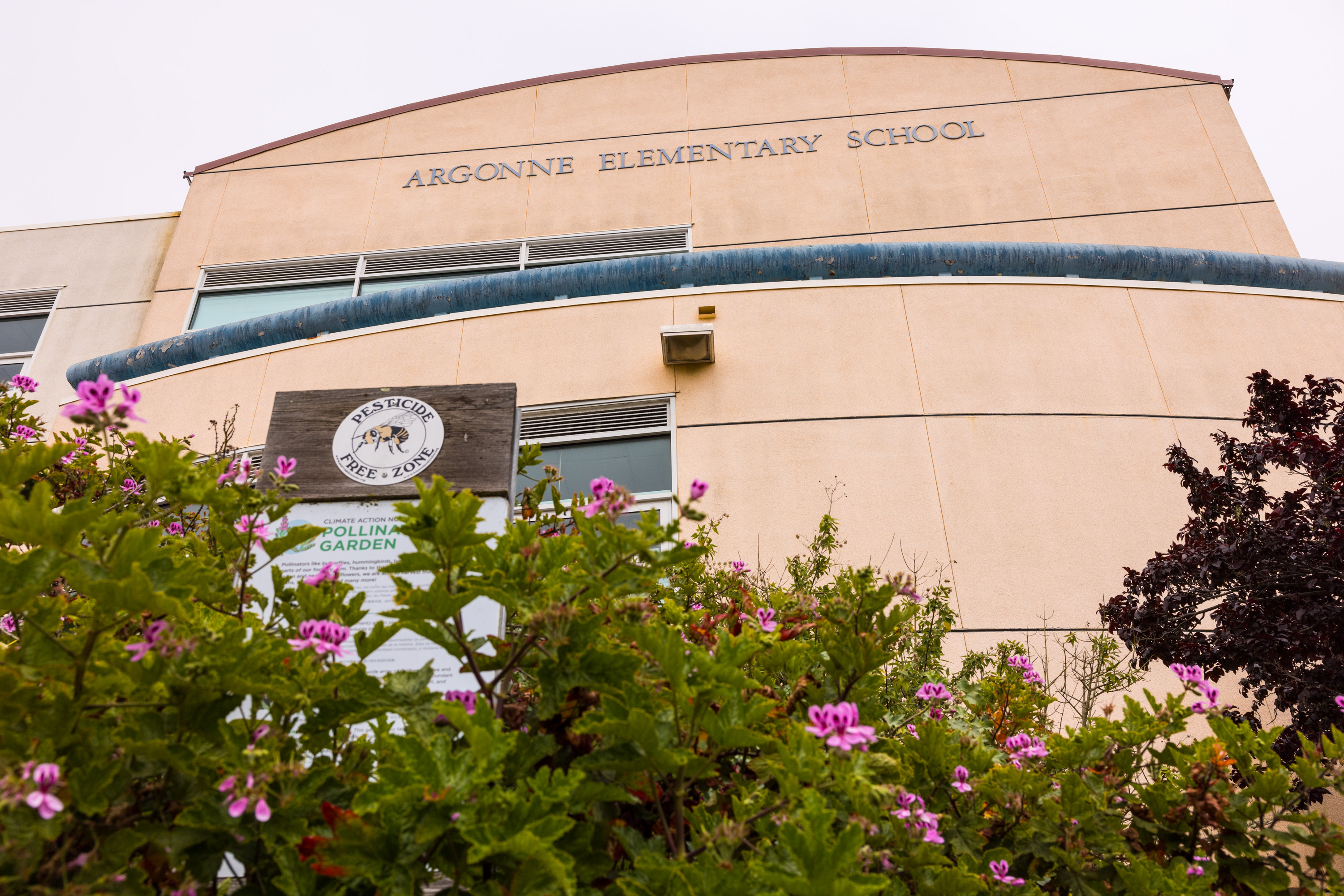 The image shows Argonne Elementary School's curved facade, with "Pesticide Free Zone" and garden signs, framed by blooming pink flowers and greenery.