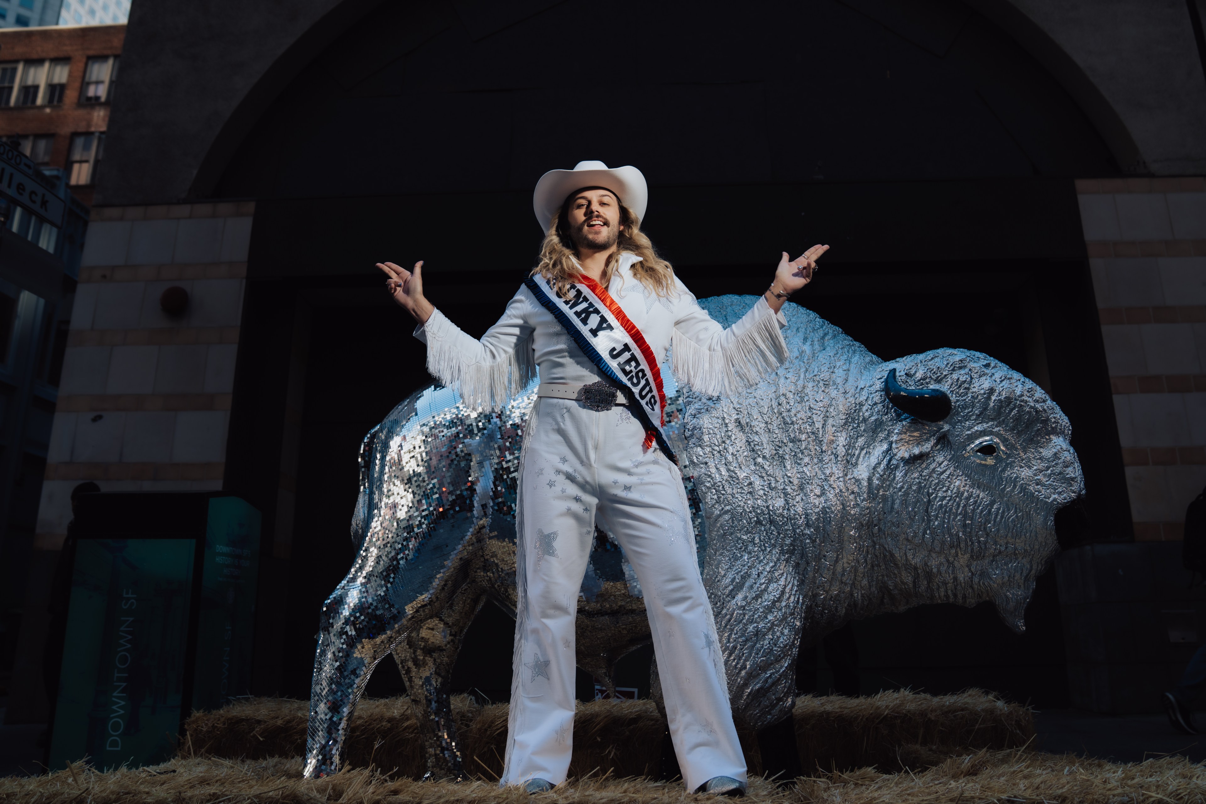 A person in a white outfit with fringe and a cowboy hat stands confidently in front of a shiny, mirrored bison statue, on a bed of hay.