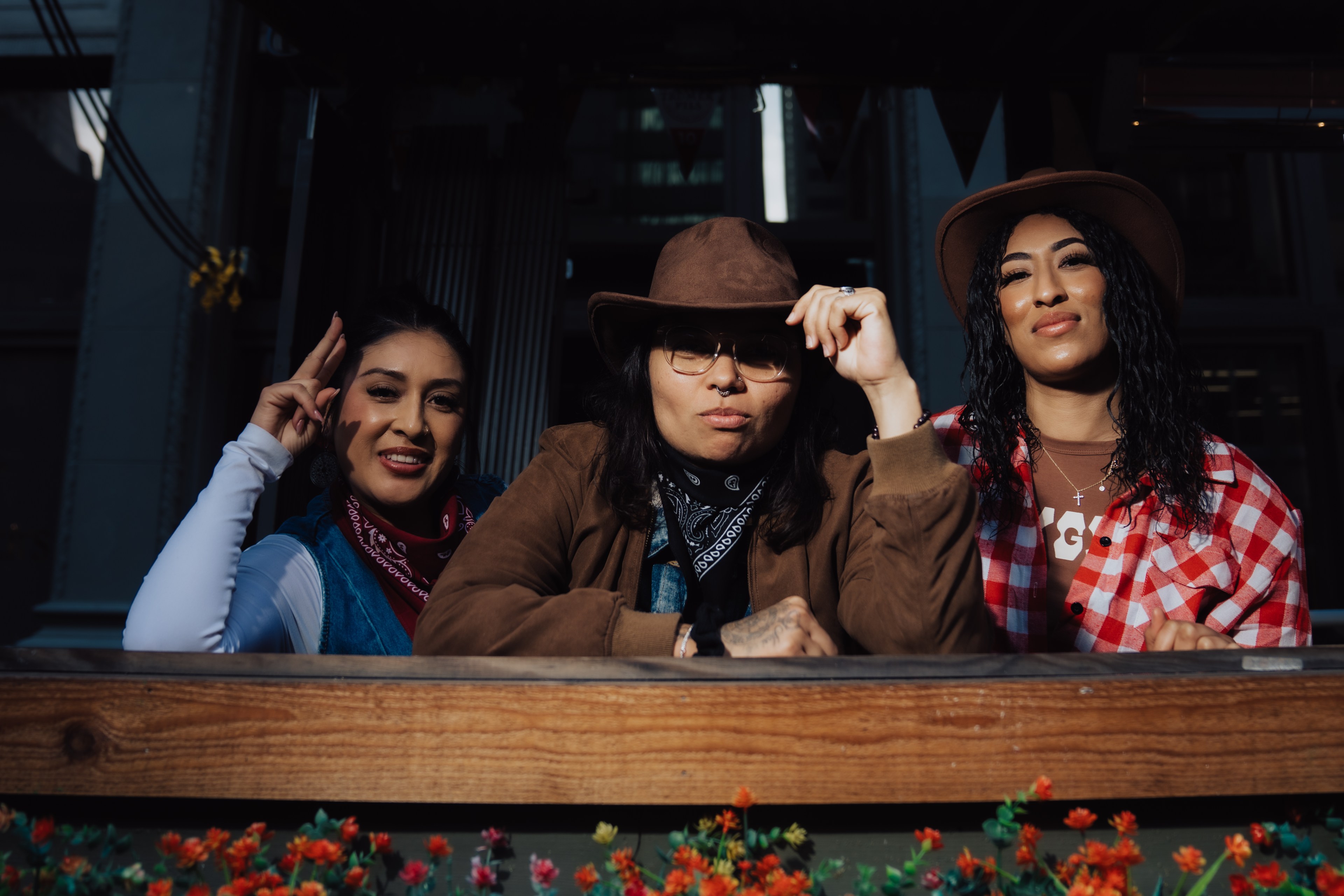 Three people are leaning over a wooden railing decorated with small flowers. They wear hats and bandanas, smiling confidently in the sunlight.