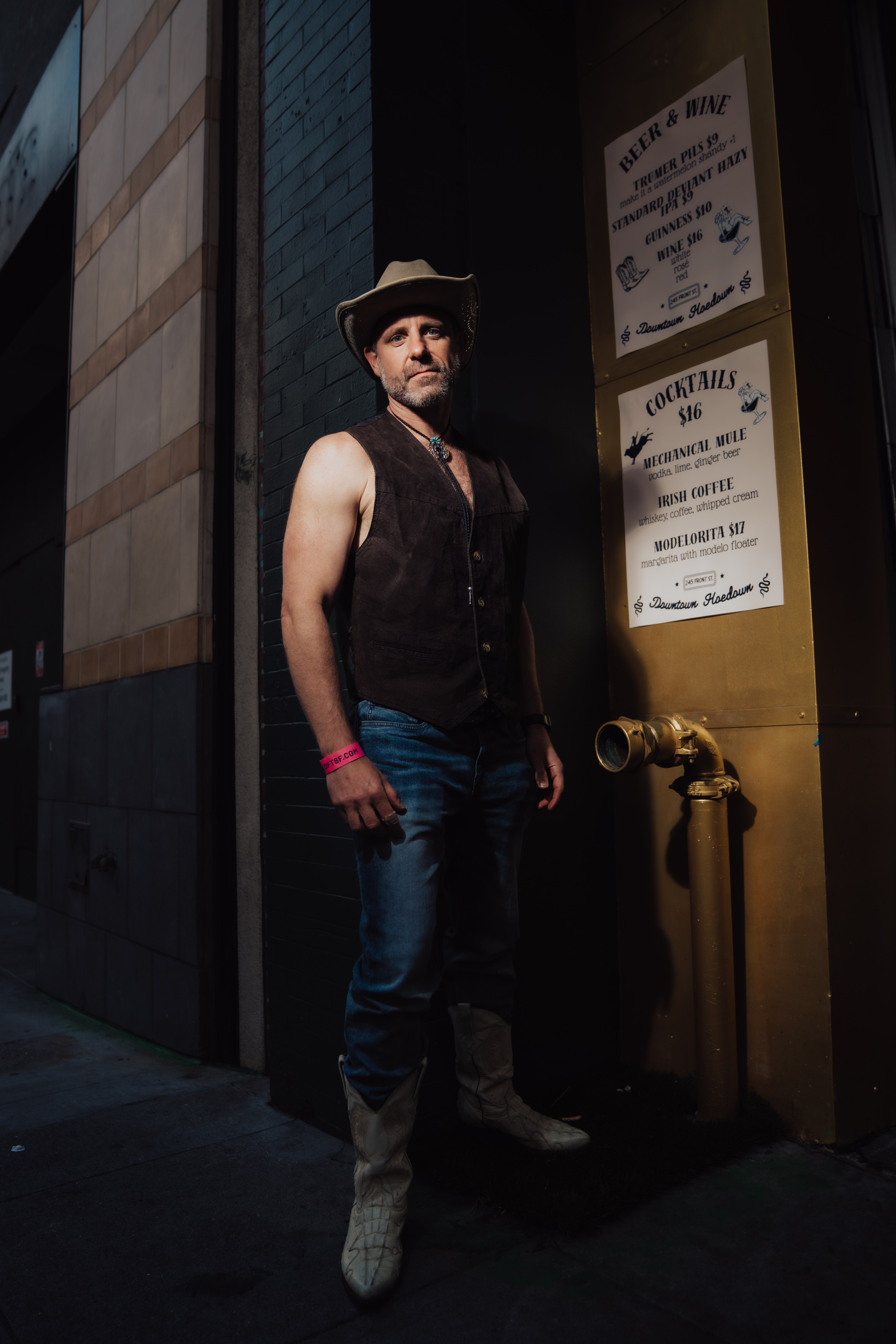 A man in a cowboy hat and sleeveless vest stands in an alley. He wears jeans and boots, with signs displaying beer, wine, and cocktail options in the background.