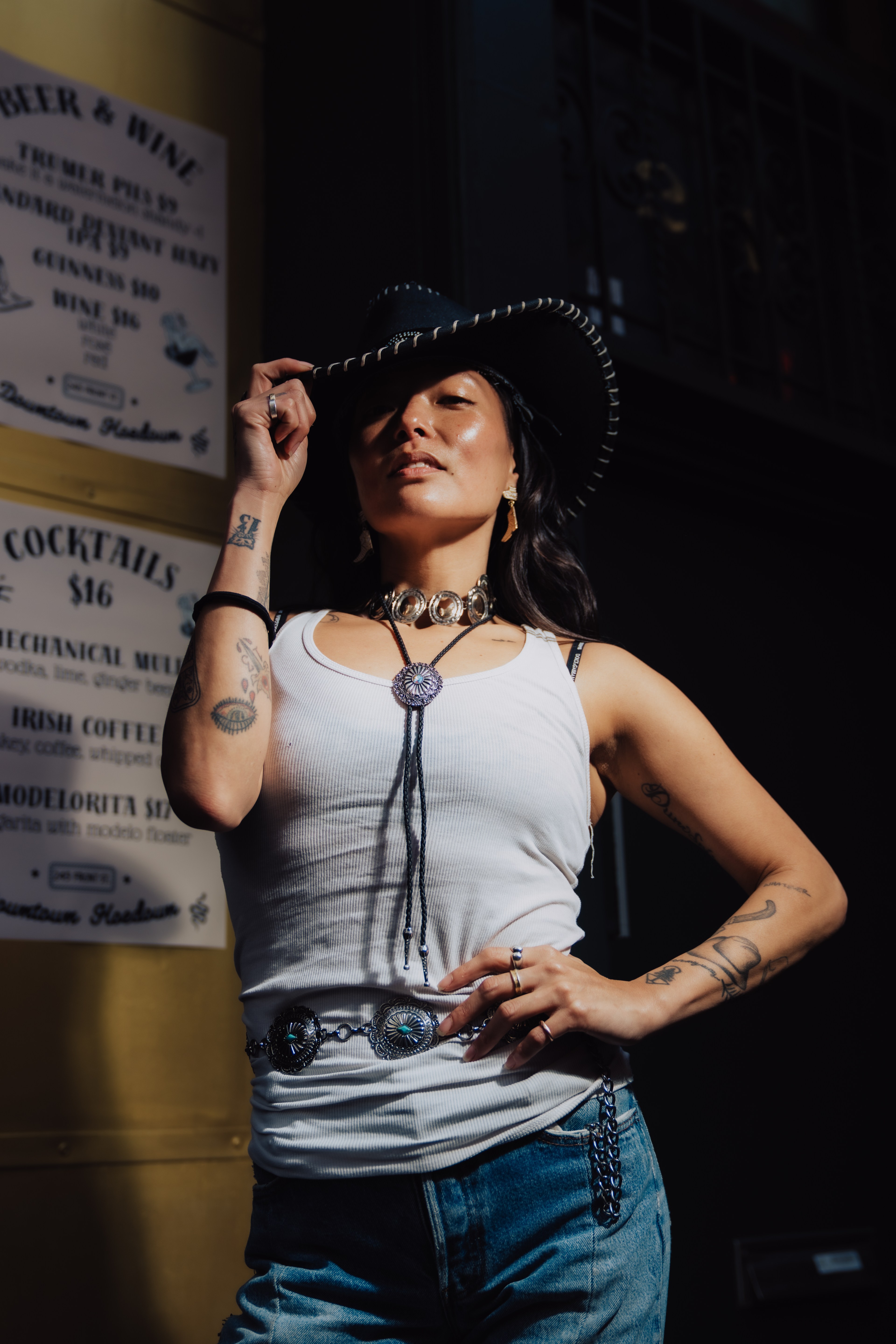A person in a cowboy hat and white tank top stands confidently, with tattoos visible on their arms. They hold the hat brim, wearing jeans and ornate jewelry.