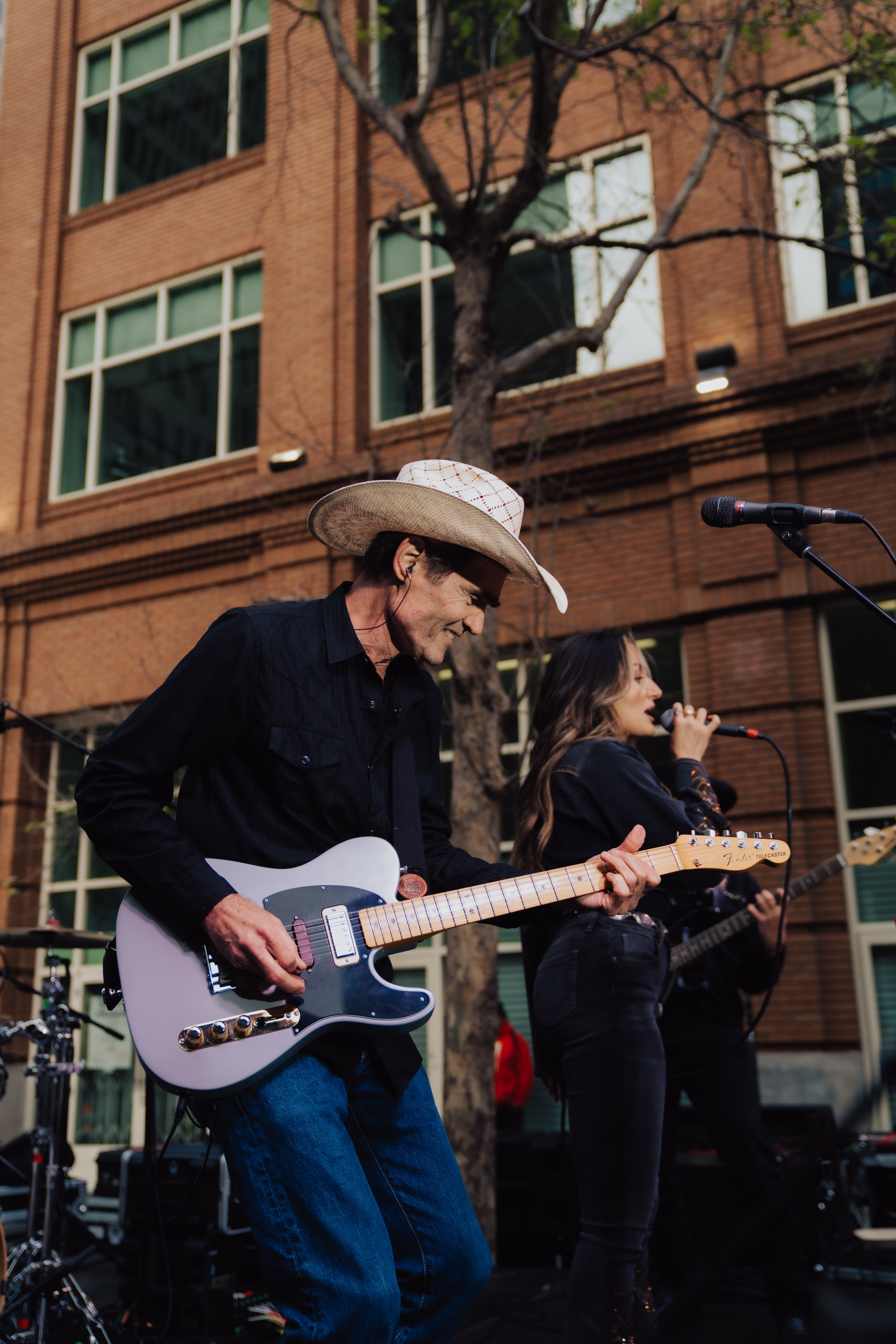 A man in a cowboy hat plays an electric guitar on an outdoor stage, while a woman sings beside him. They are in front of a brick building with large windows.