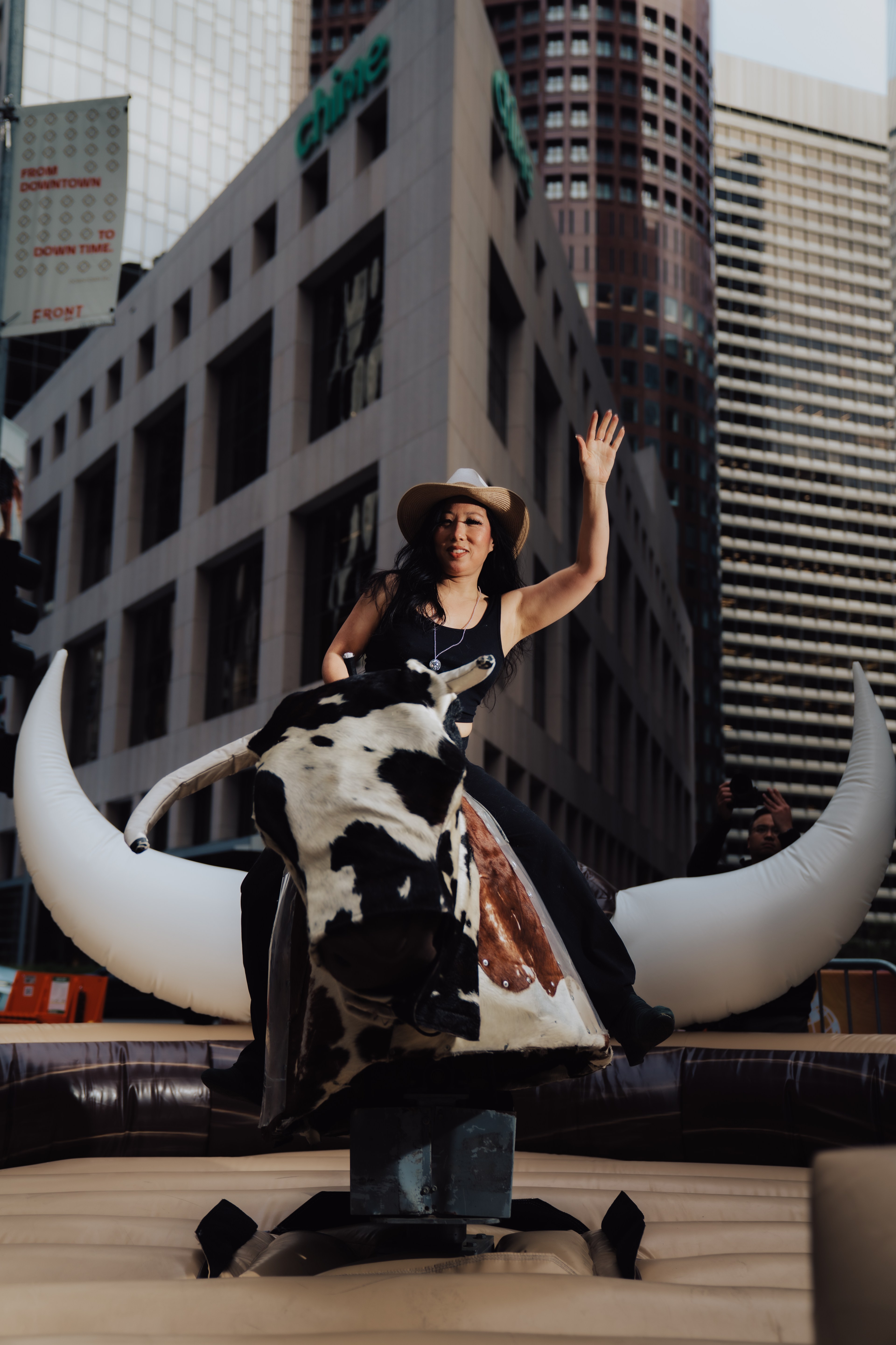 A person wearing a cowboy hat rides a mechanical bull in an urban setting, surrounded by tall buildings. They appear to be waving with one hand.