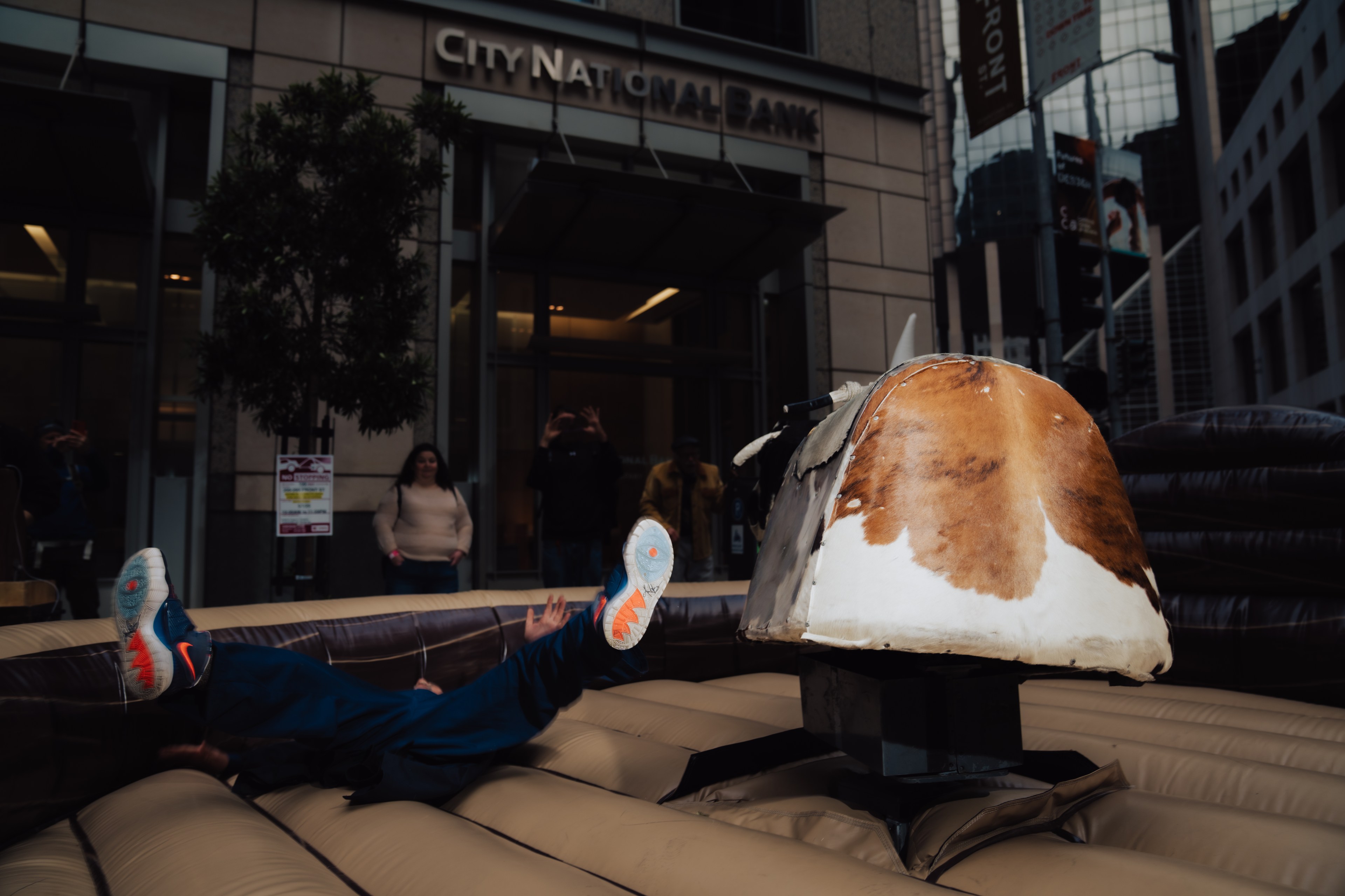 A person falls off a mechanical bull in front of a building with a &quot;City National Bank&quot; sign. Their feet are in the air as bystanders watch and take photos.