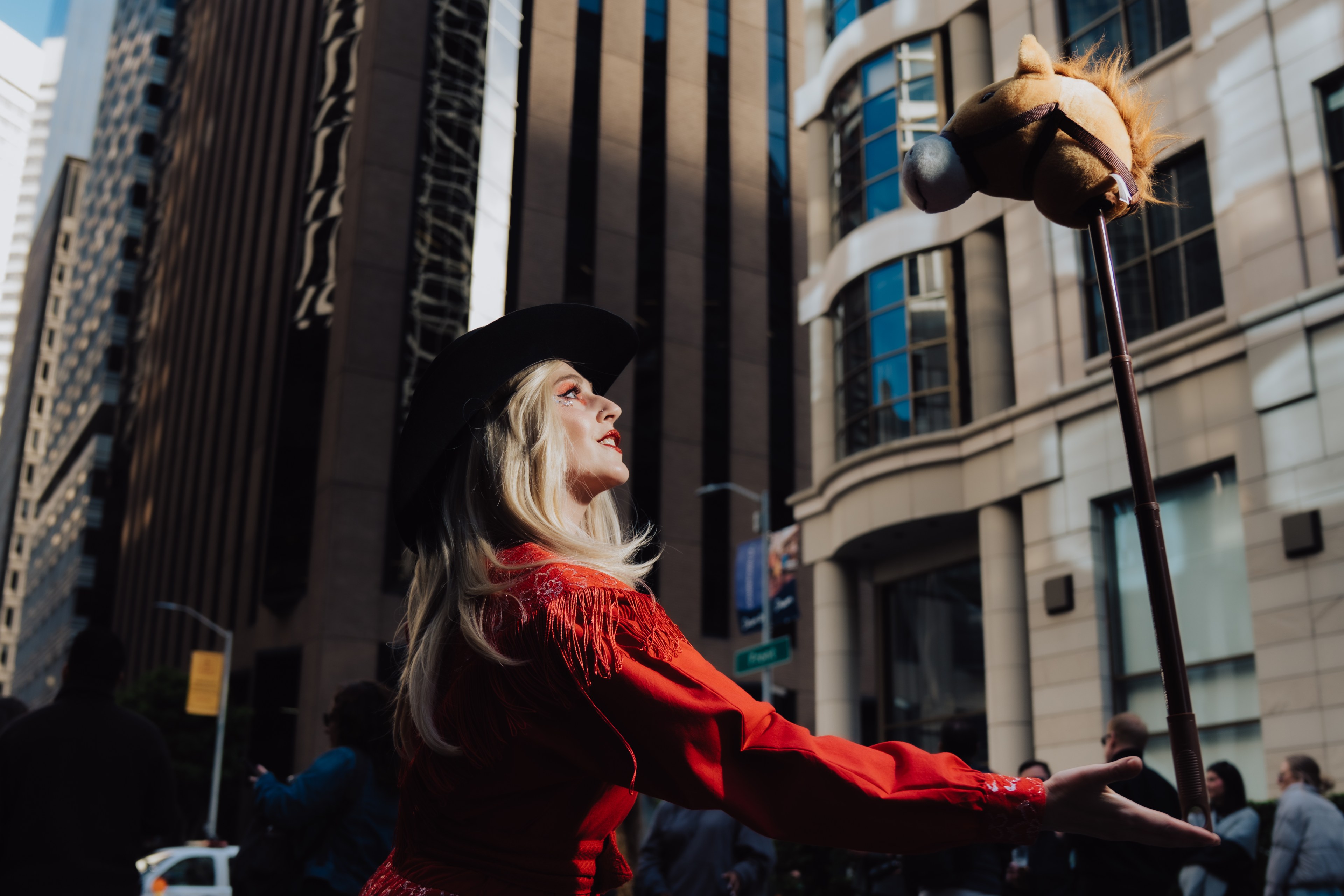 A person wearing a black hat and red outfit holds a toy horse on a stick, gazing upwards. They stand in an urban area with tall buildings around.