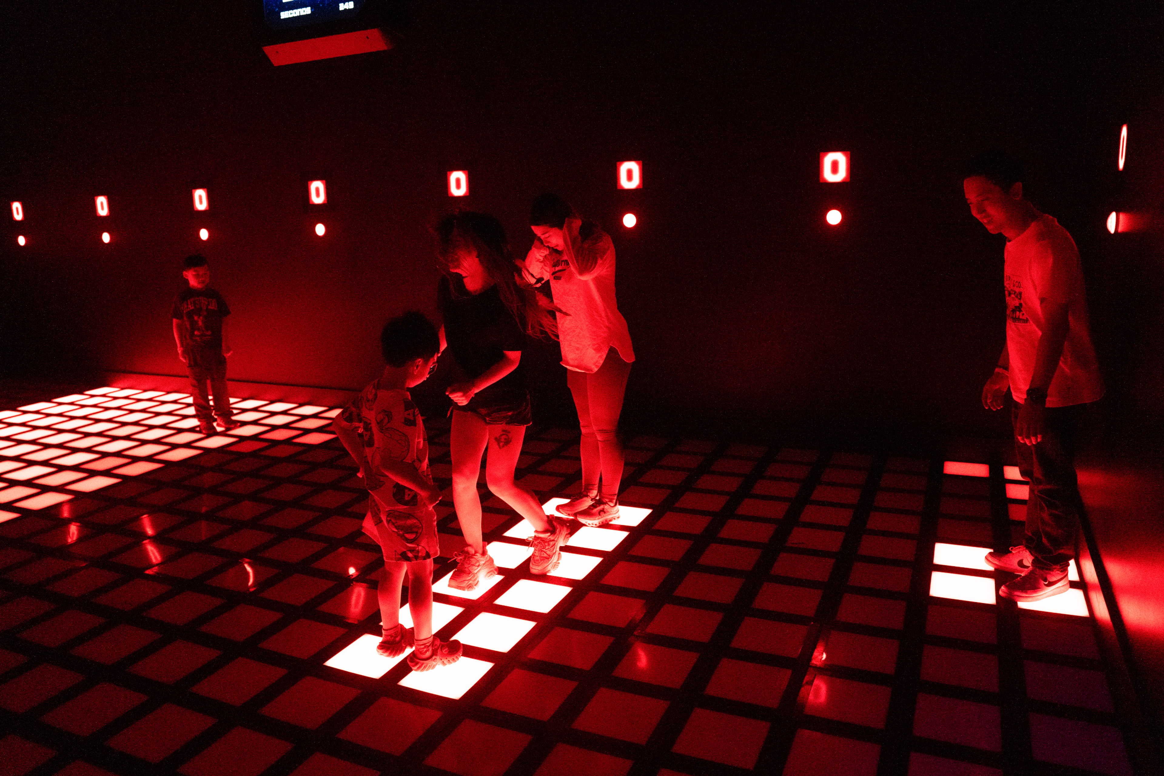 The image shows people standing on a glowing red grid floor in a dark room. The walls have small red lights and digital zeros displayed.