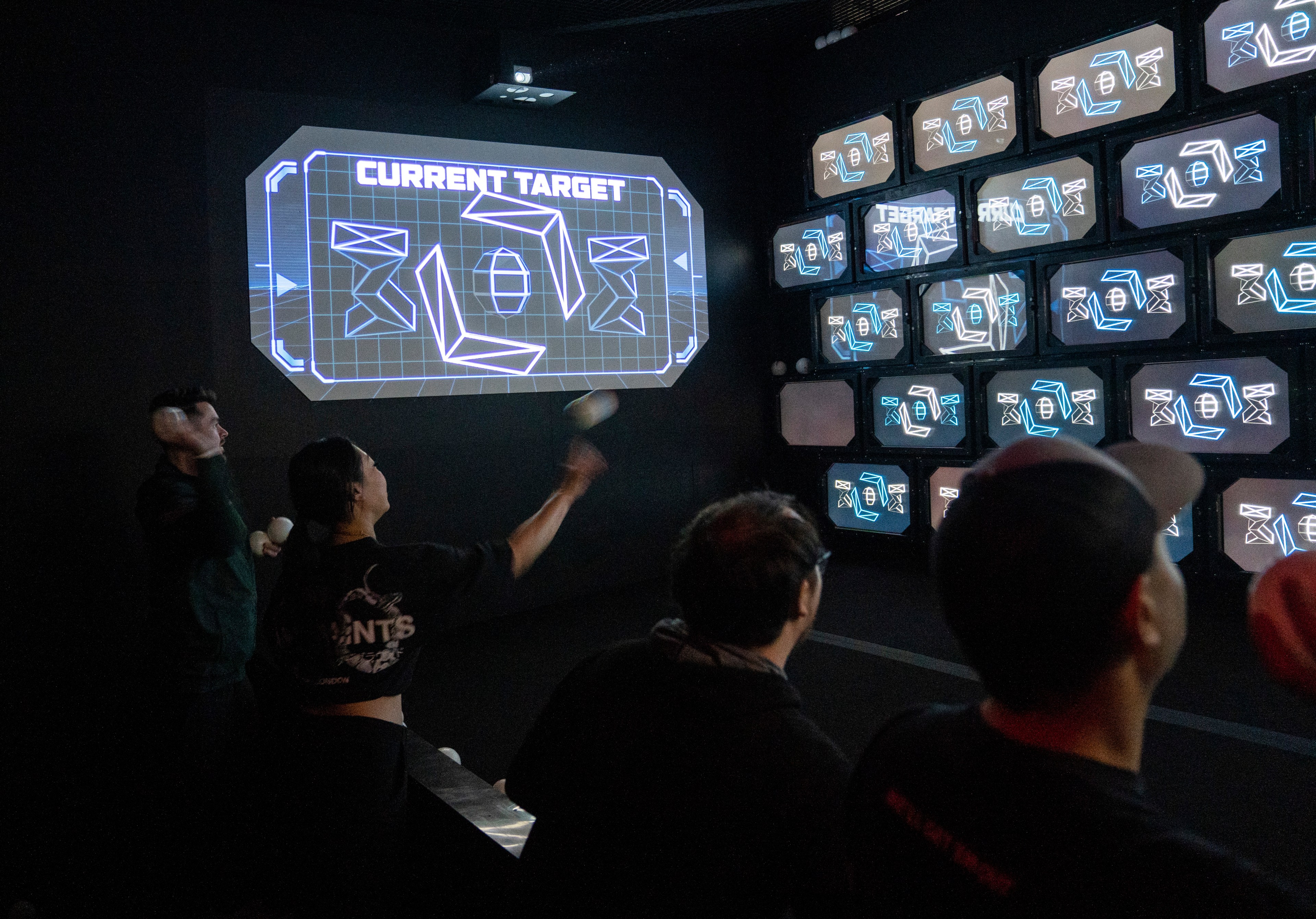 A group of people is throwing balls at a wall of screens displaying geometric shapes. The main screen reads "CURRENT TARGET," indicating a game or activity.