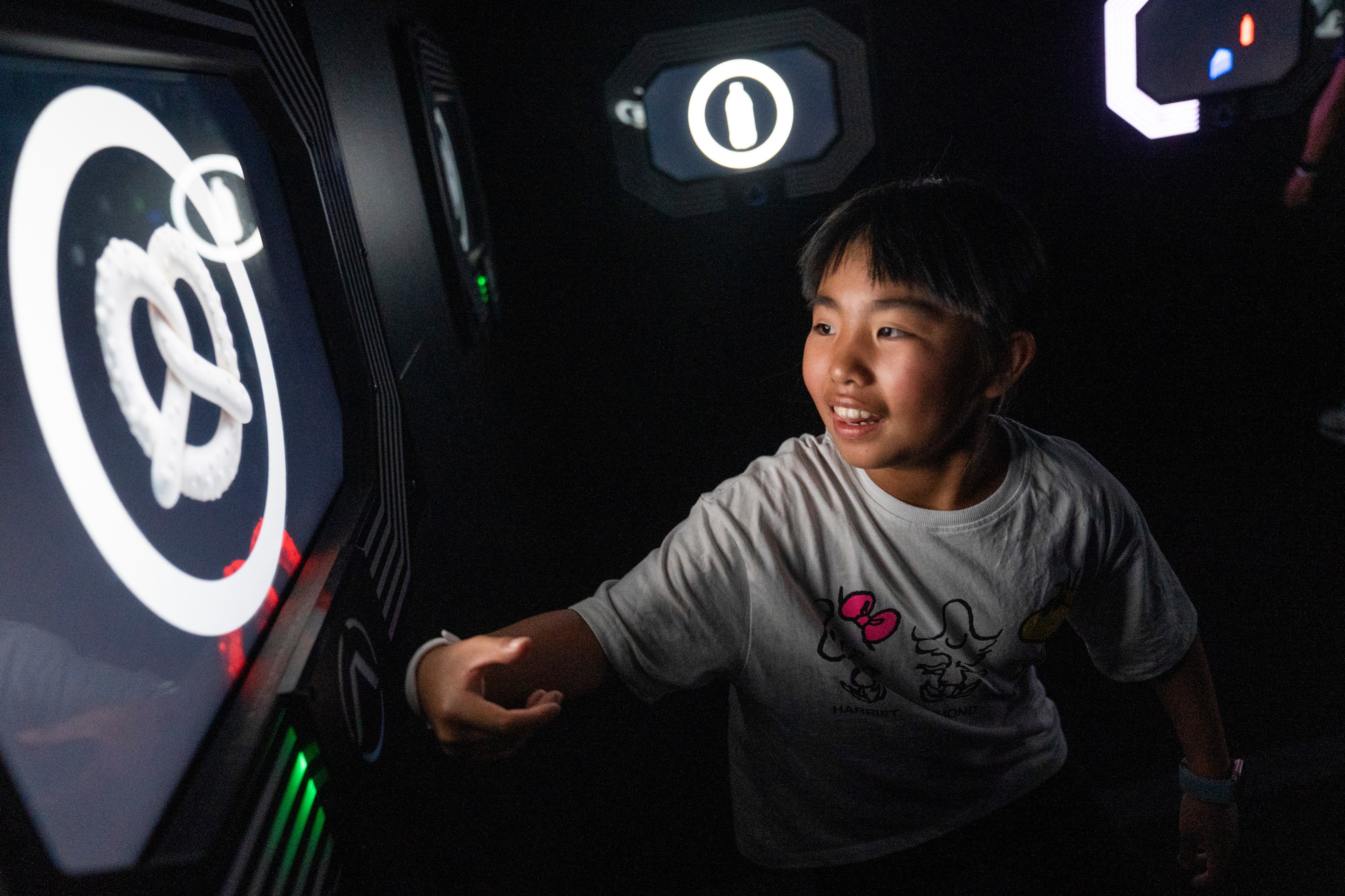 A child interacts with a touch screen showing a pretzel inside a circle, on a wall with illuminated symbols. They look engaged and curious.