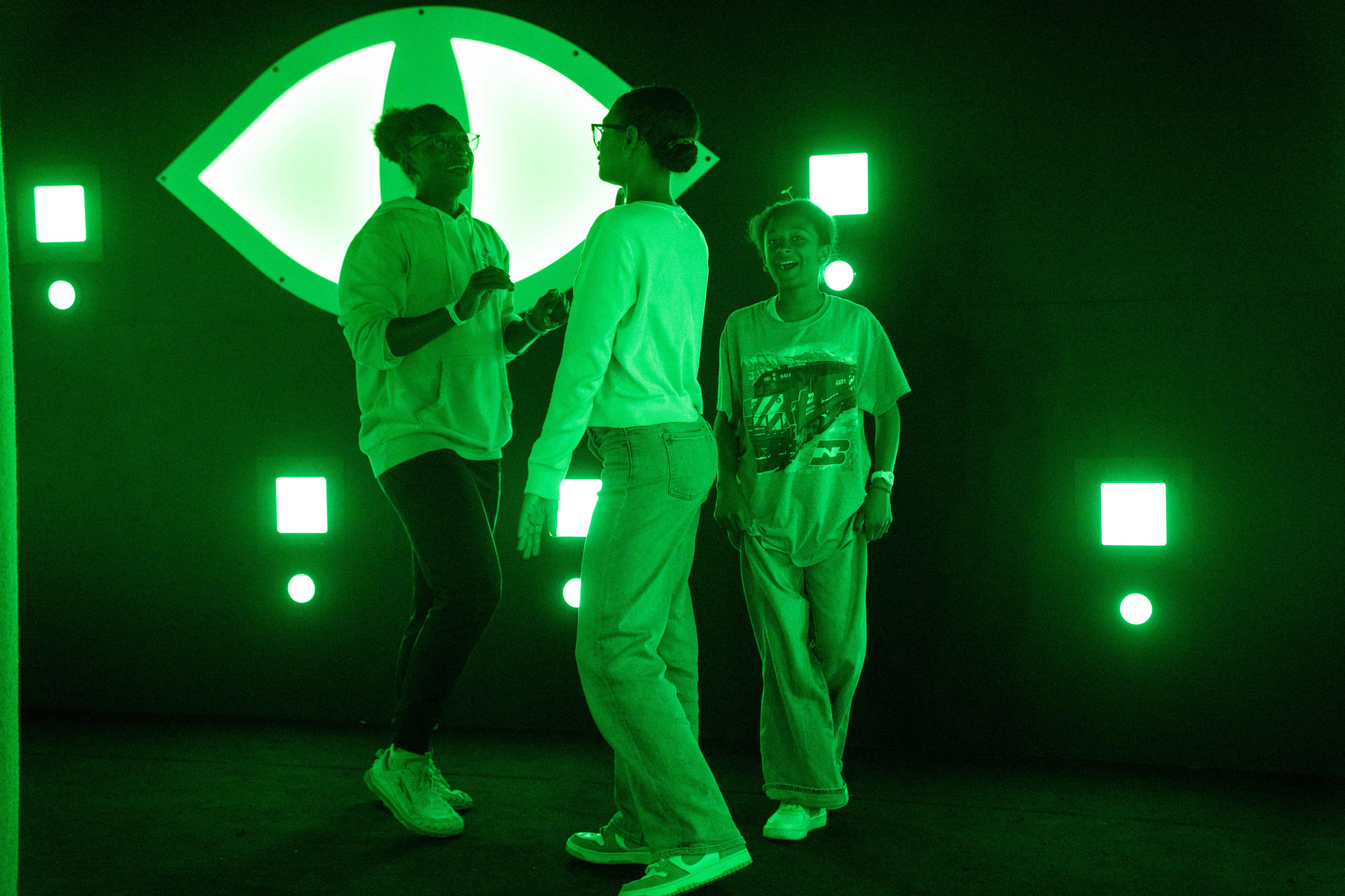 Three people are smiling and interacting in a dark room illuminated by green neon lights and geometric shapes on the walls.