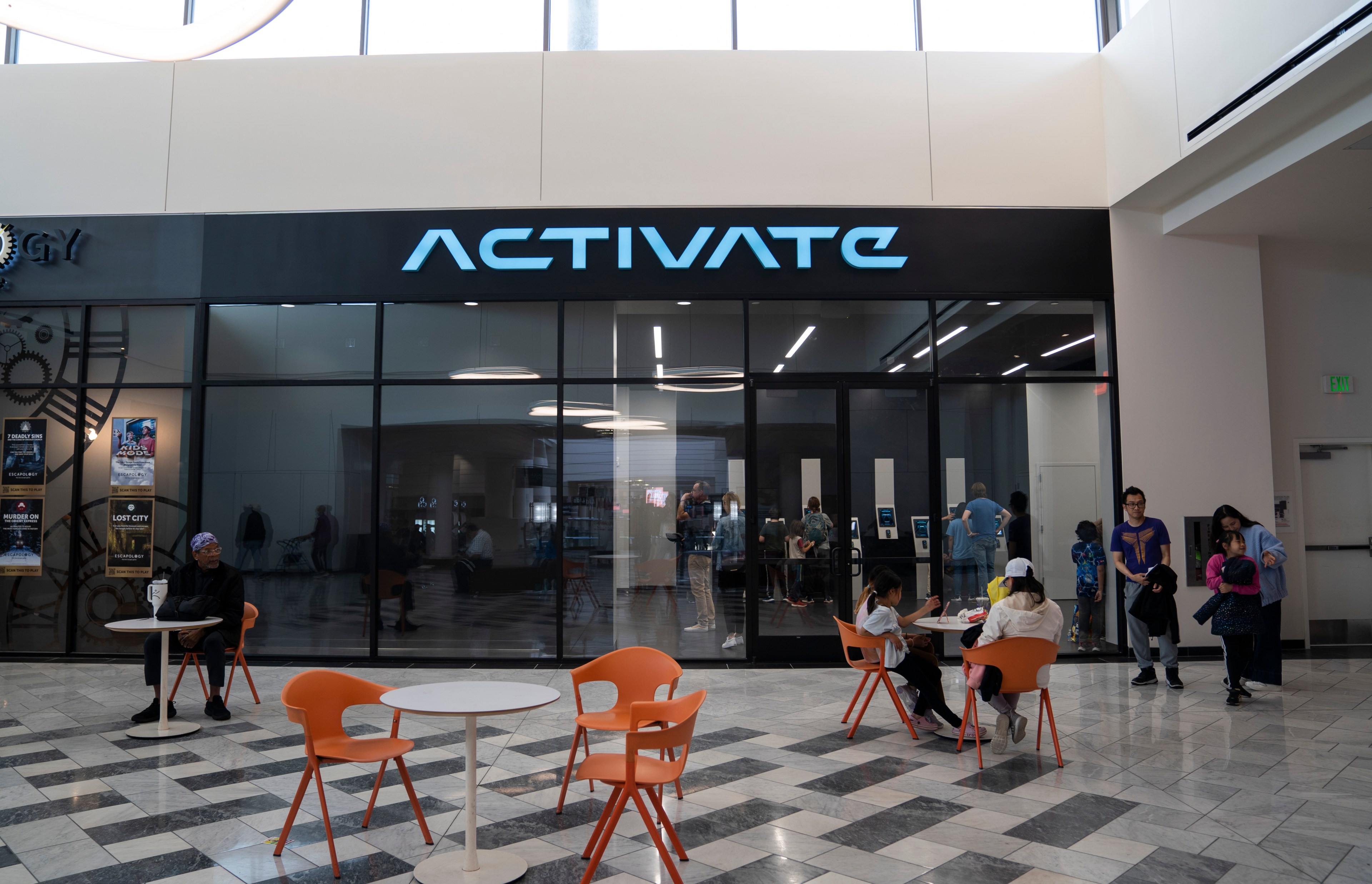 The image shows a store called &quot;ACTIVATE&quot; with a glass front, bright interior lighting, and people inside. In front, there are orange chairs and tables with people sitting.