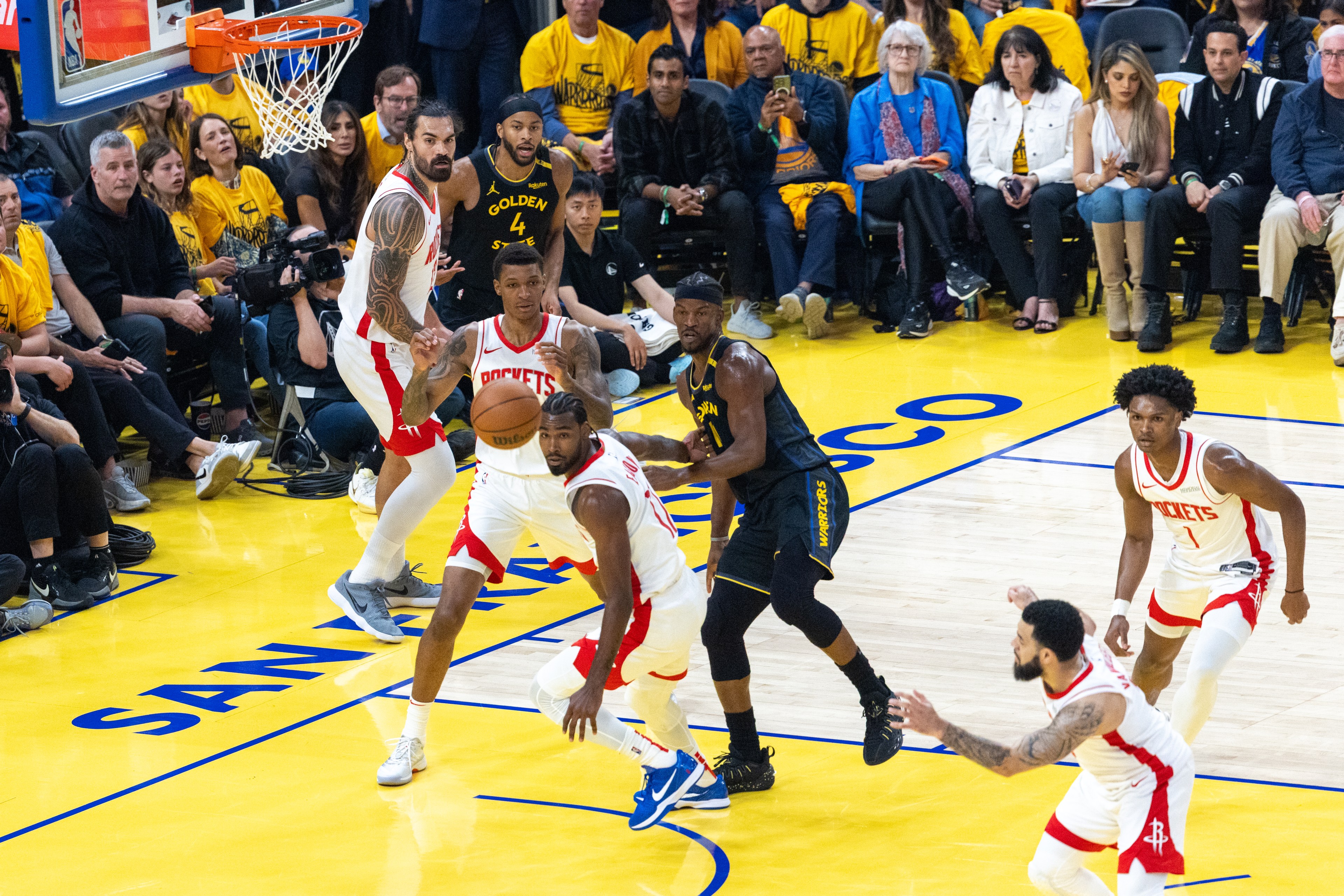 Basketball players from teams in red and black jerseys are in action on a court. One player holds the ball, while others guard and position themselves.