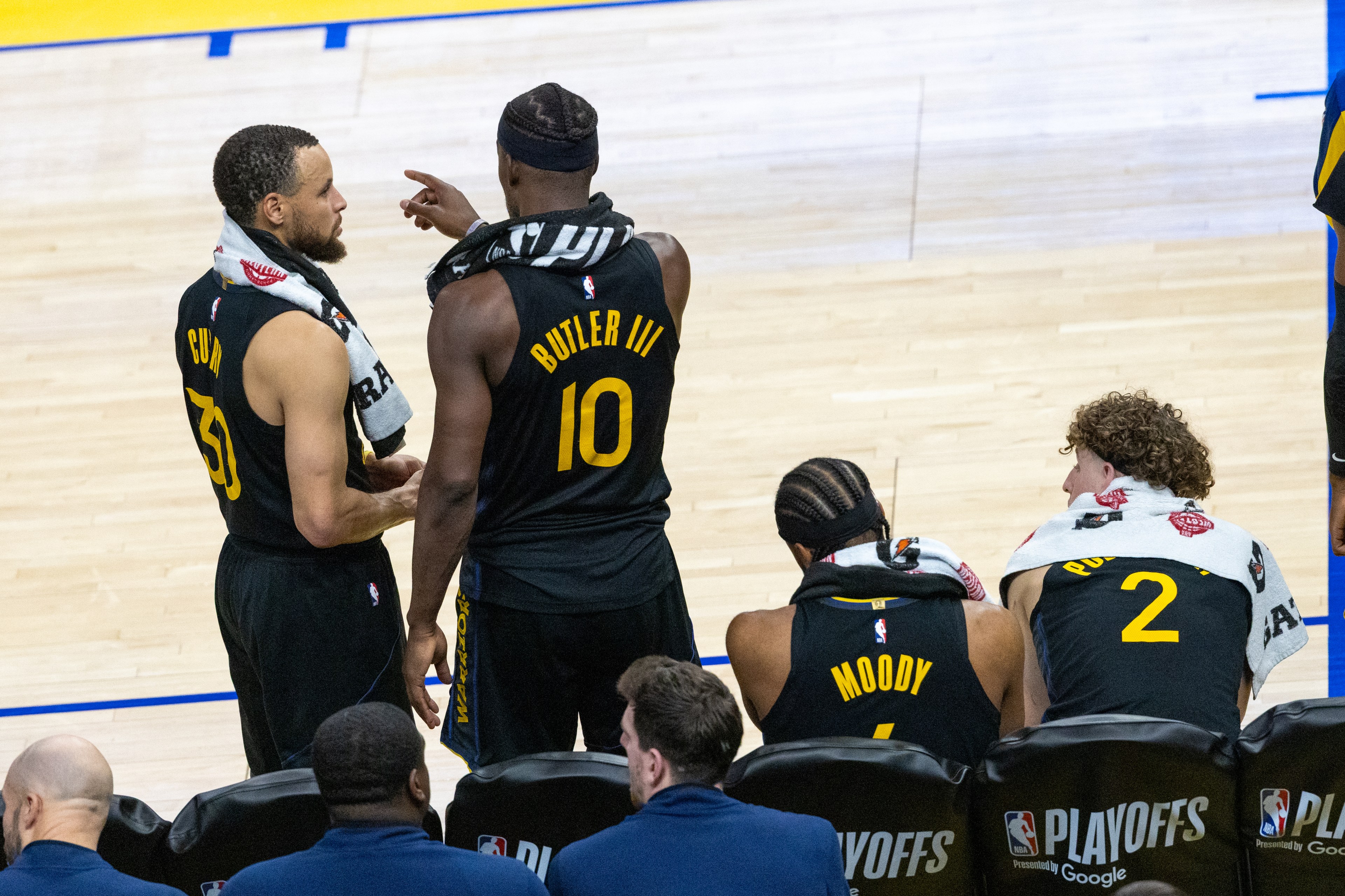 Two basketball players stand talking, one pointing. Two others sit on the bench with towels around their necks, and "PLAYOFFS" is visible on the seats.