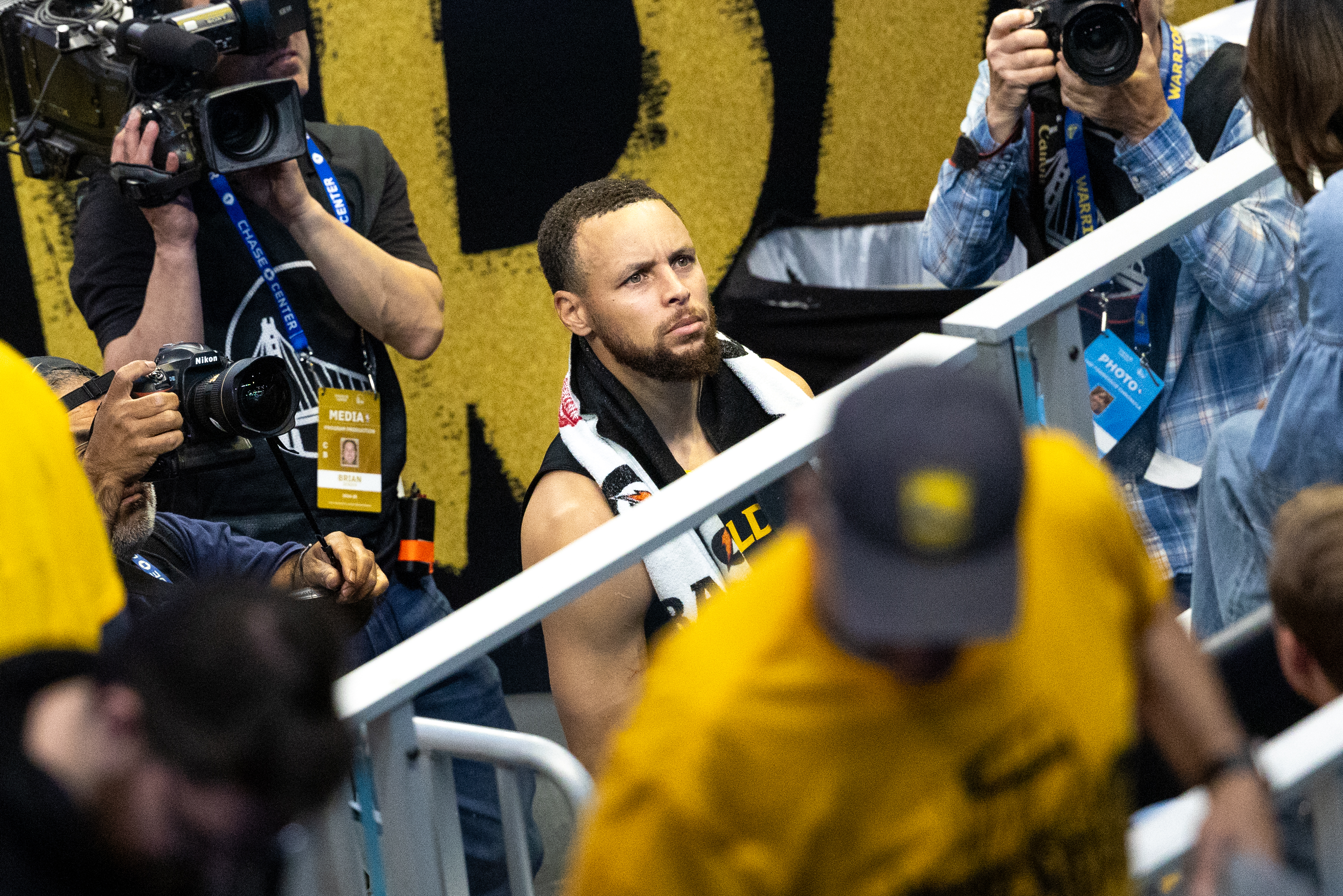 A focused athlete with a towel around his neck is captured by multiple photographers and camera operators in a bustling, media-heavy environment.