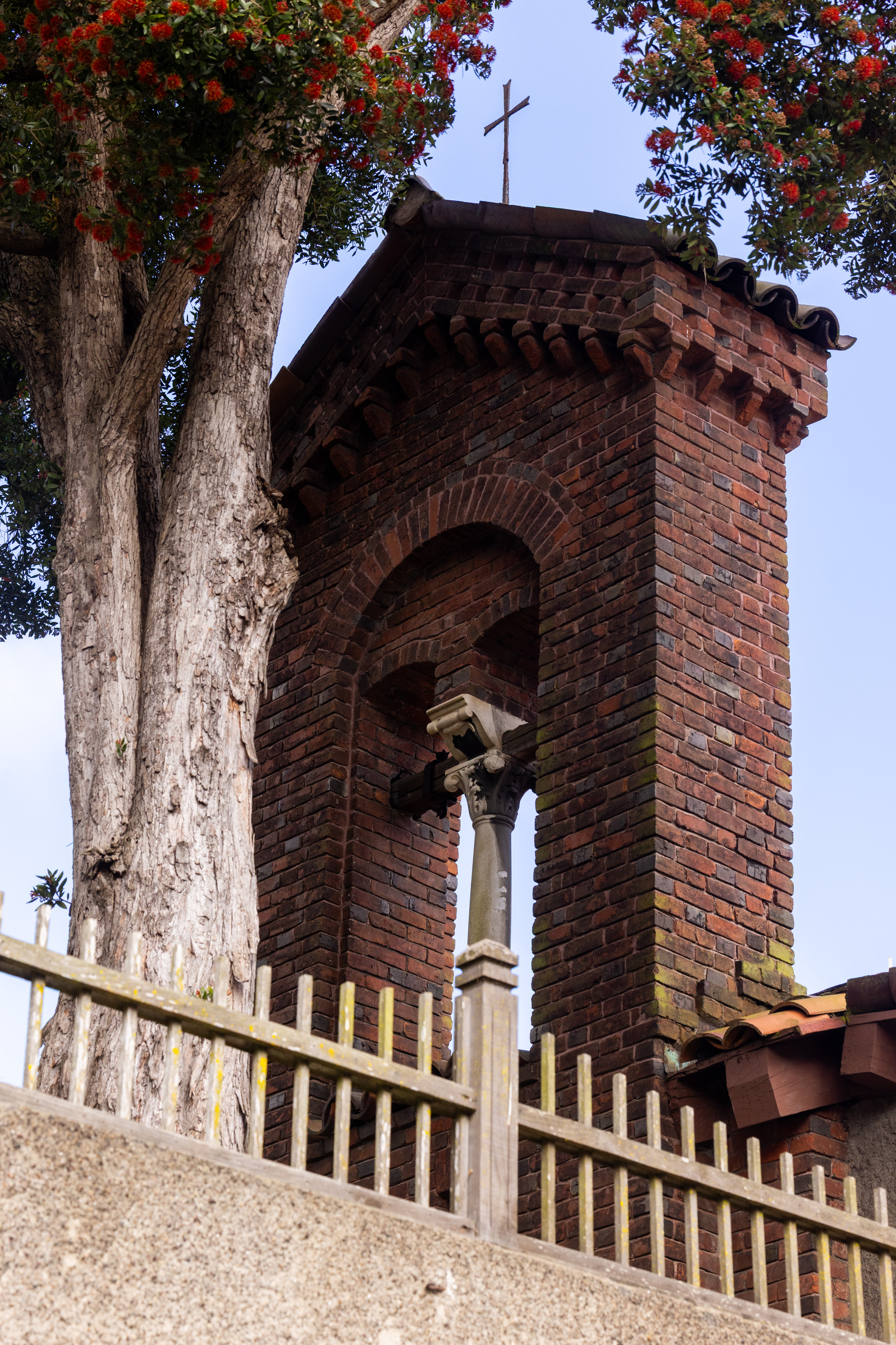 A brick church tower with a cross on top is framed by a tall tree with red flowers. A wooden fence is visible in the foreground.