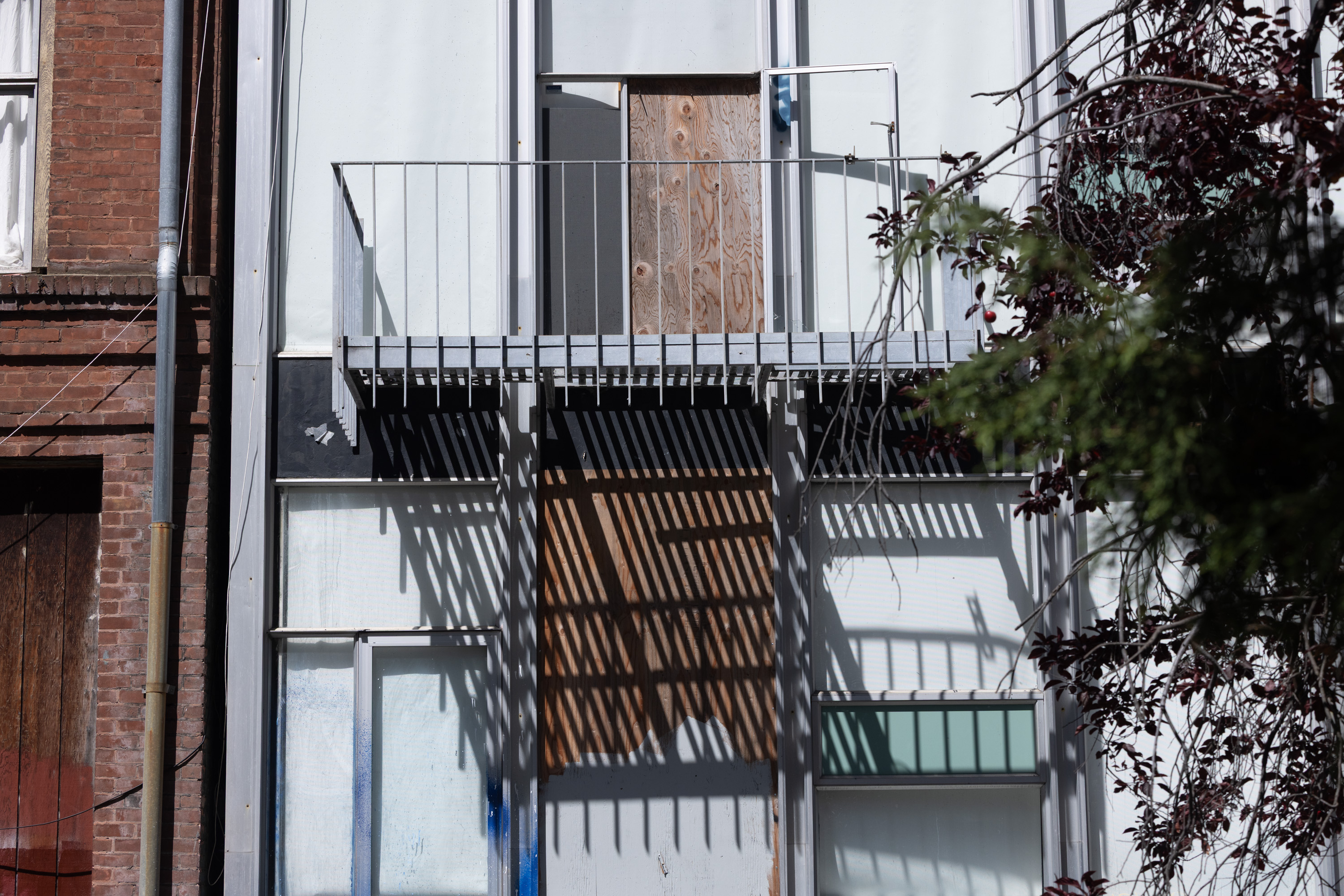 The image shows an old building facade with a metal balcony casting striped shadows on boarded-up white and glass panels, surrounded by trees on the right.