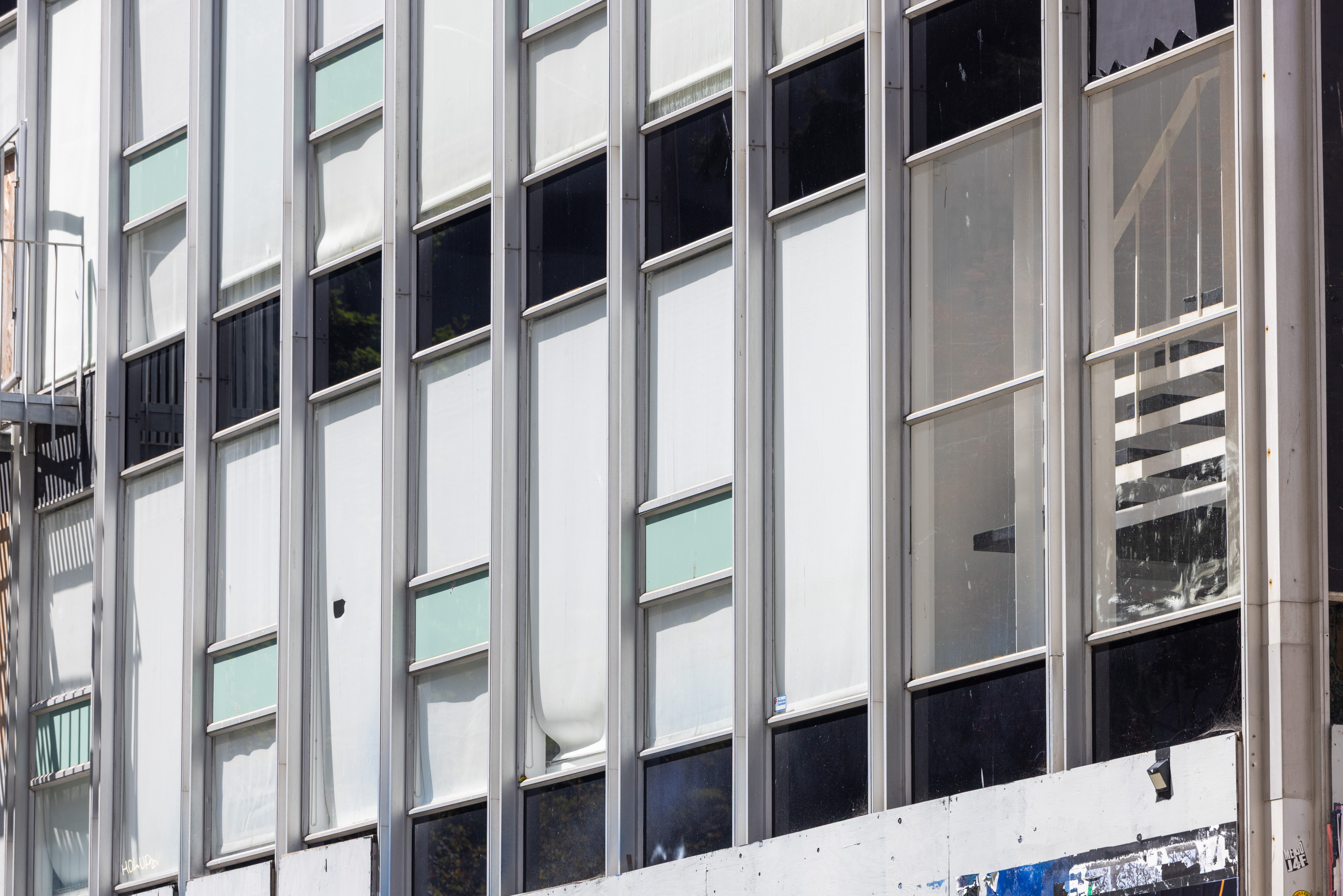 The image shows a building facade with large vertical windows framed in metal. Some windows have blinds partially drawn, and a fire escape stairs are visible on the left.