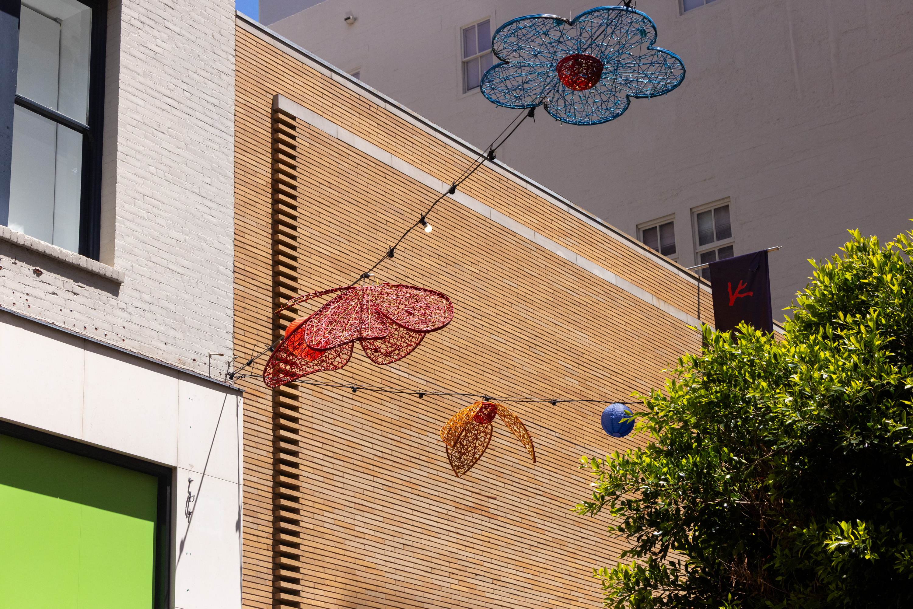 Colorful wire sculptures shaped like flowers hang above a street, set against brick and white walls, with a green window and some trees visible.