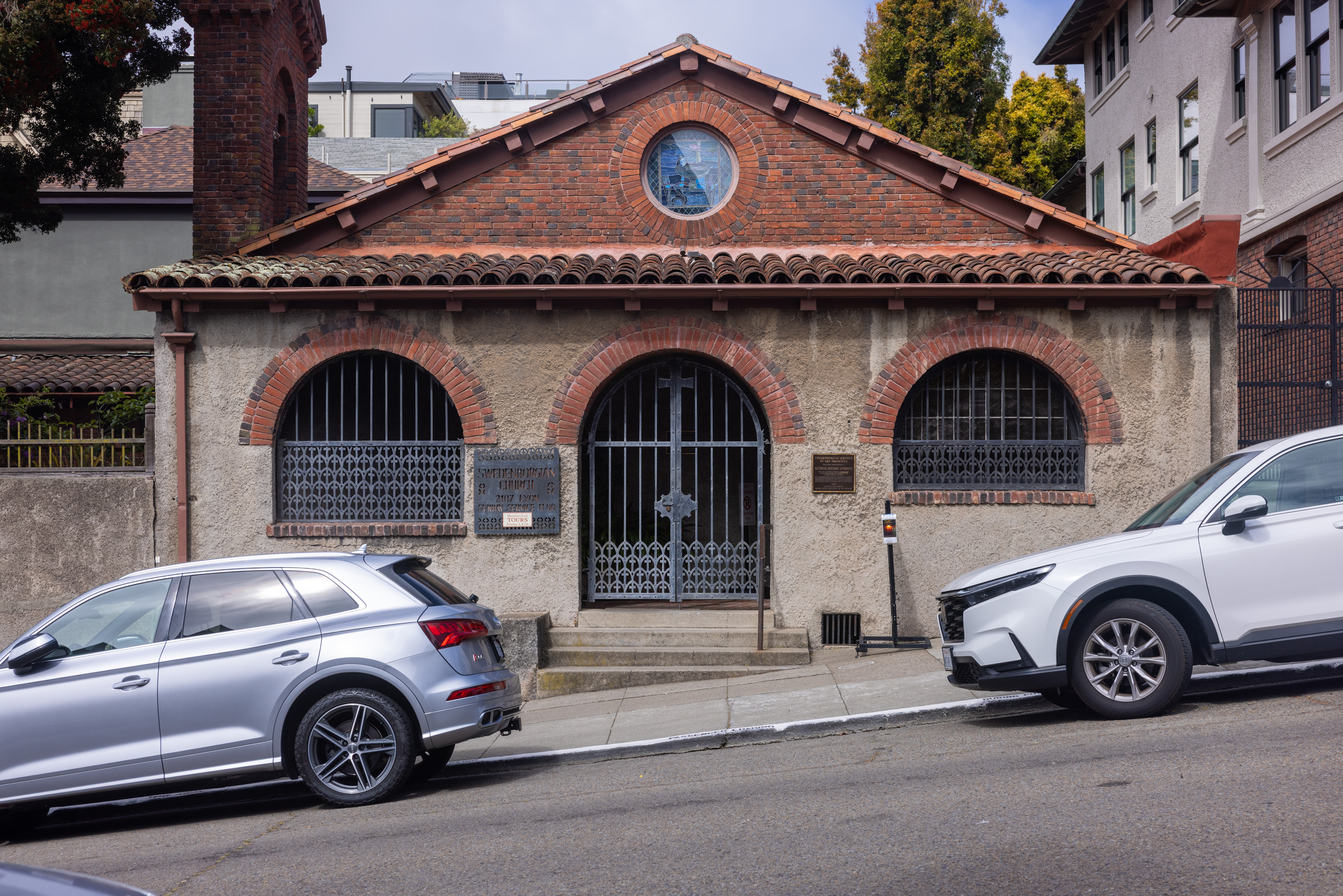 A small, rustic building with a red tile roof and arched windows is situated on a sloped street, flanked by two parked cars.