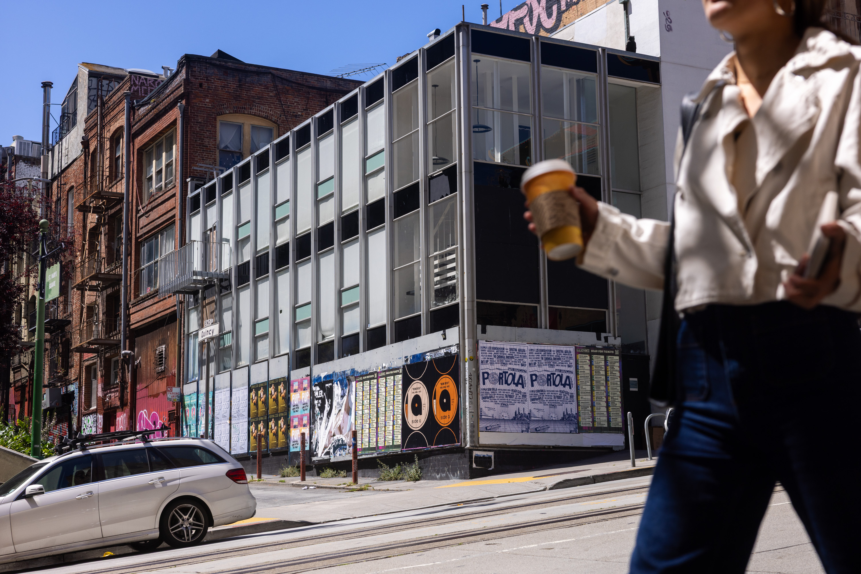 A woman holding a coffee cup walks past a modern glass building with posters on it. Old brick buildings line the street, and a white car is parked nearby.