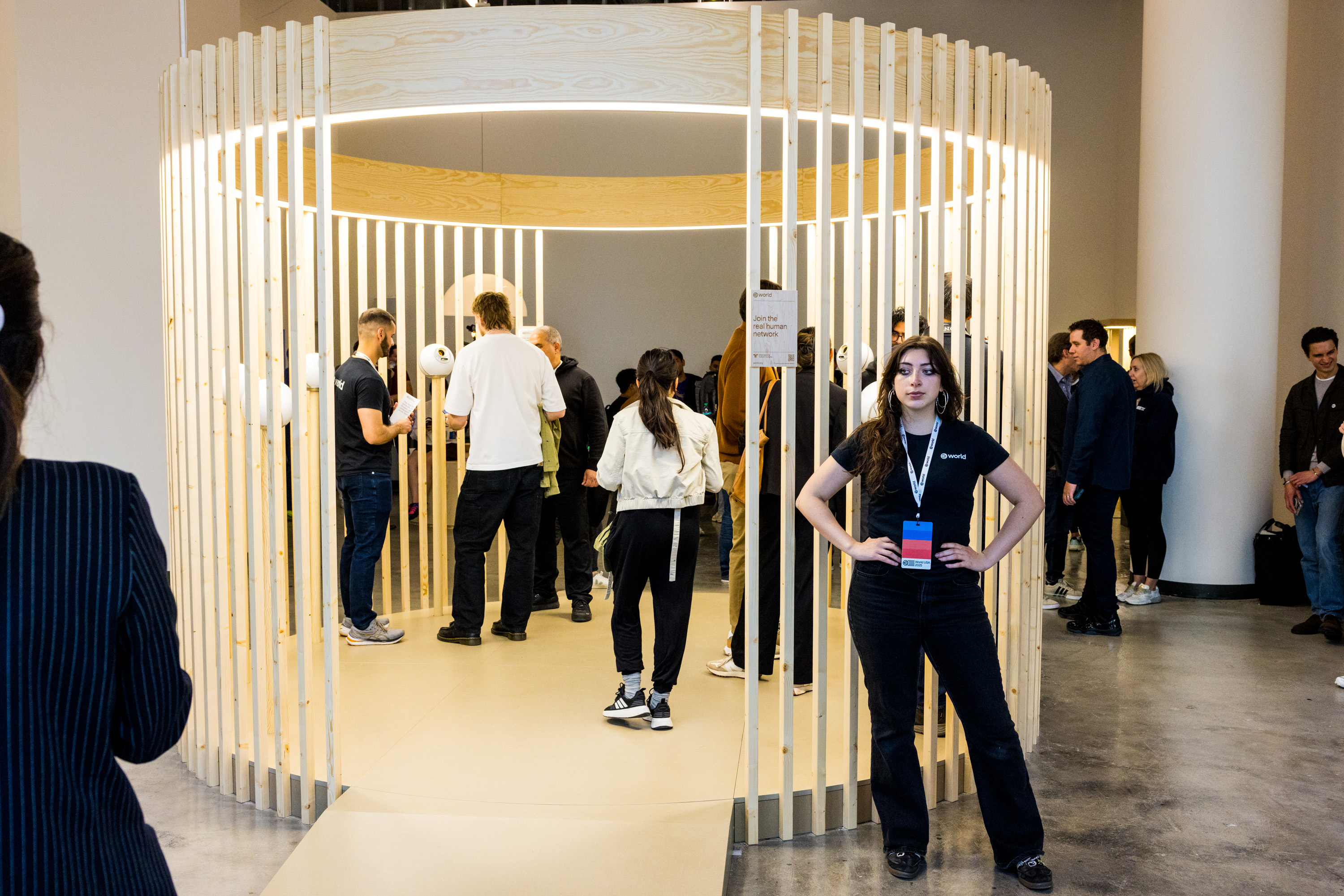The image shows a modern, circular wooden installation with people gathered inside and outside. A person stands at the entrance looking attentive, wearing a black shirt.