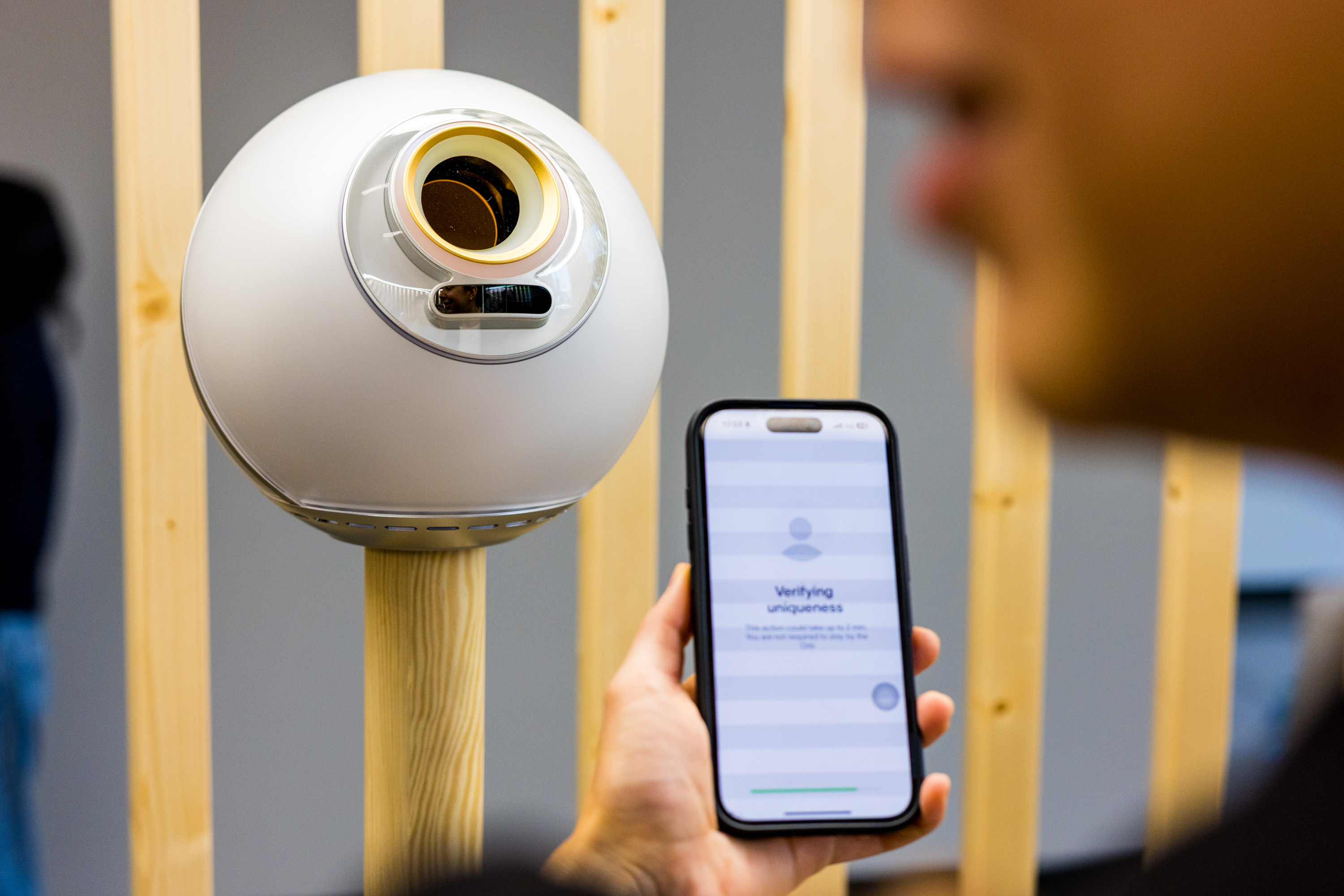 A person holds a smartphone displaying &quot;Verifying uniqueness&quot; next to a futuristic, spherical device mounted on a wooden stand.