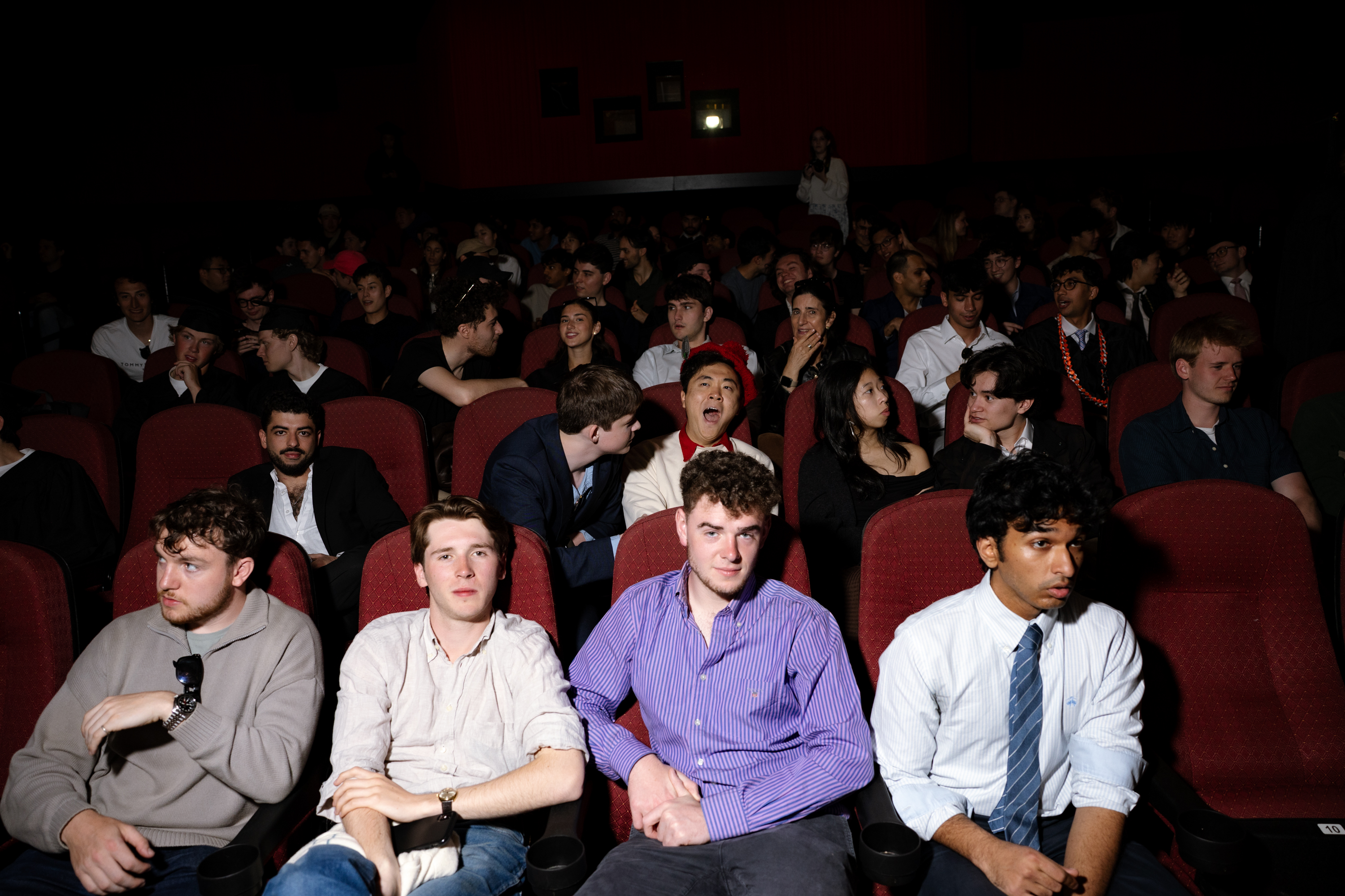 A dim theater filled with people sitting in rows of red seats, some chatting and others looking around. The atmosphere is lively and informal.