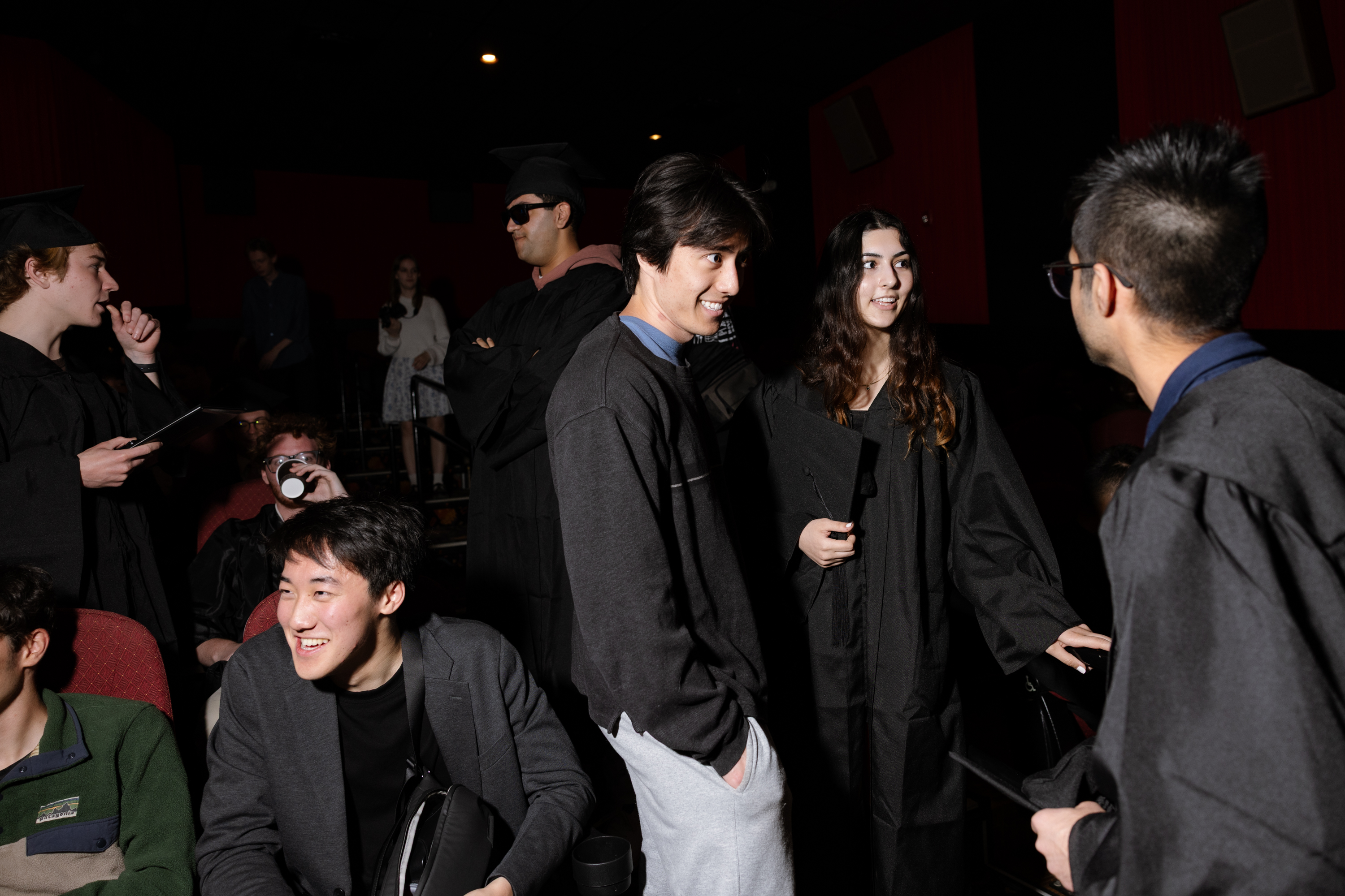 A group of young adults, some in graduation gowns and caps, chat and smile in a dimly lit room. One person drinks from a cup, and others hold diplomas.
