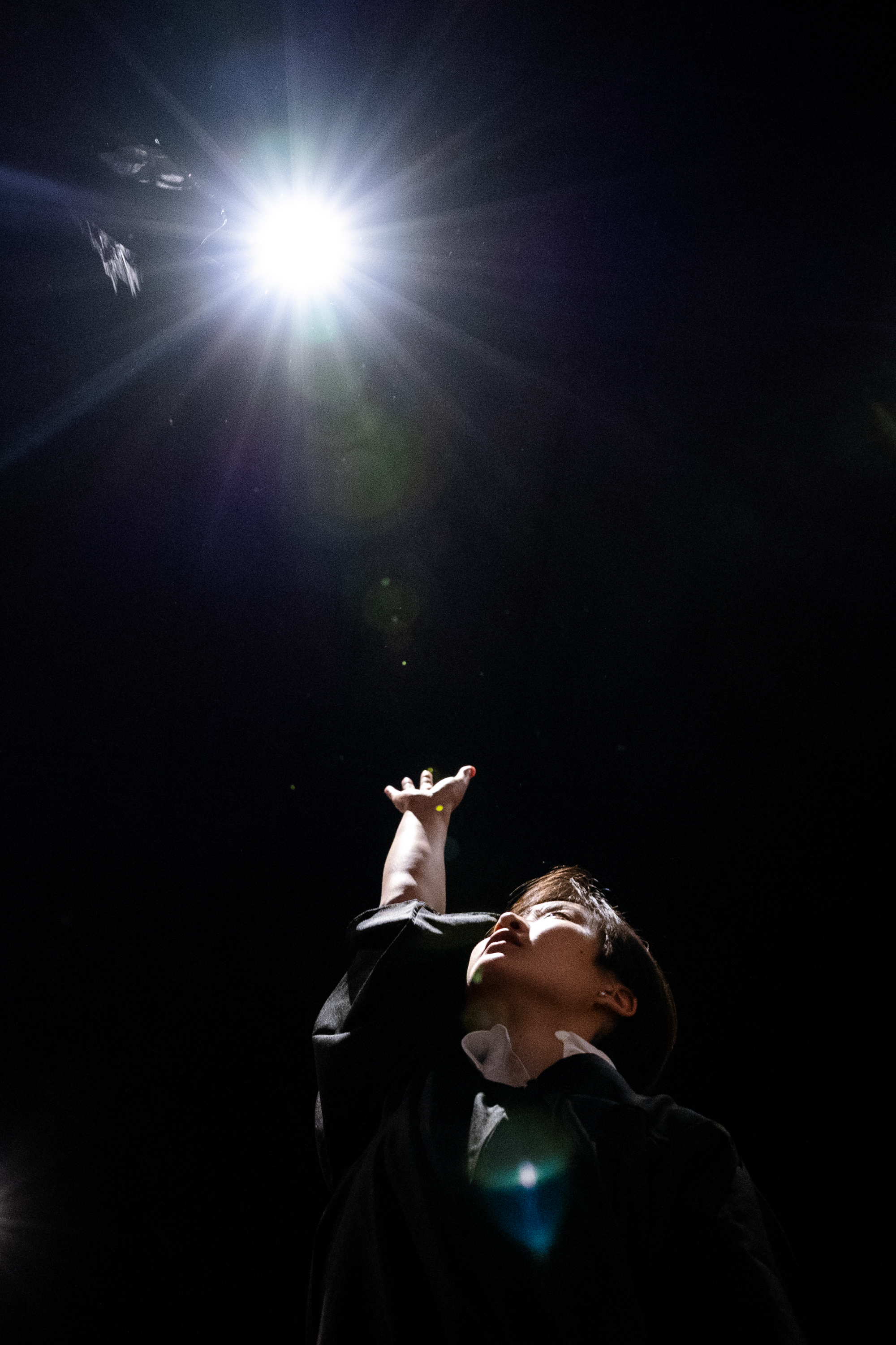 A person is reaching up toward a bright light above them, set against a dark background. Their face is illuminated slightly by the light.