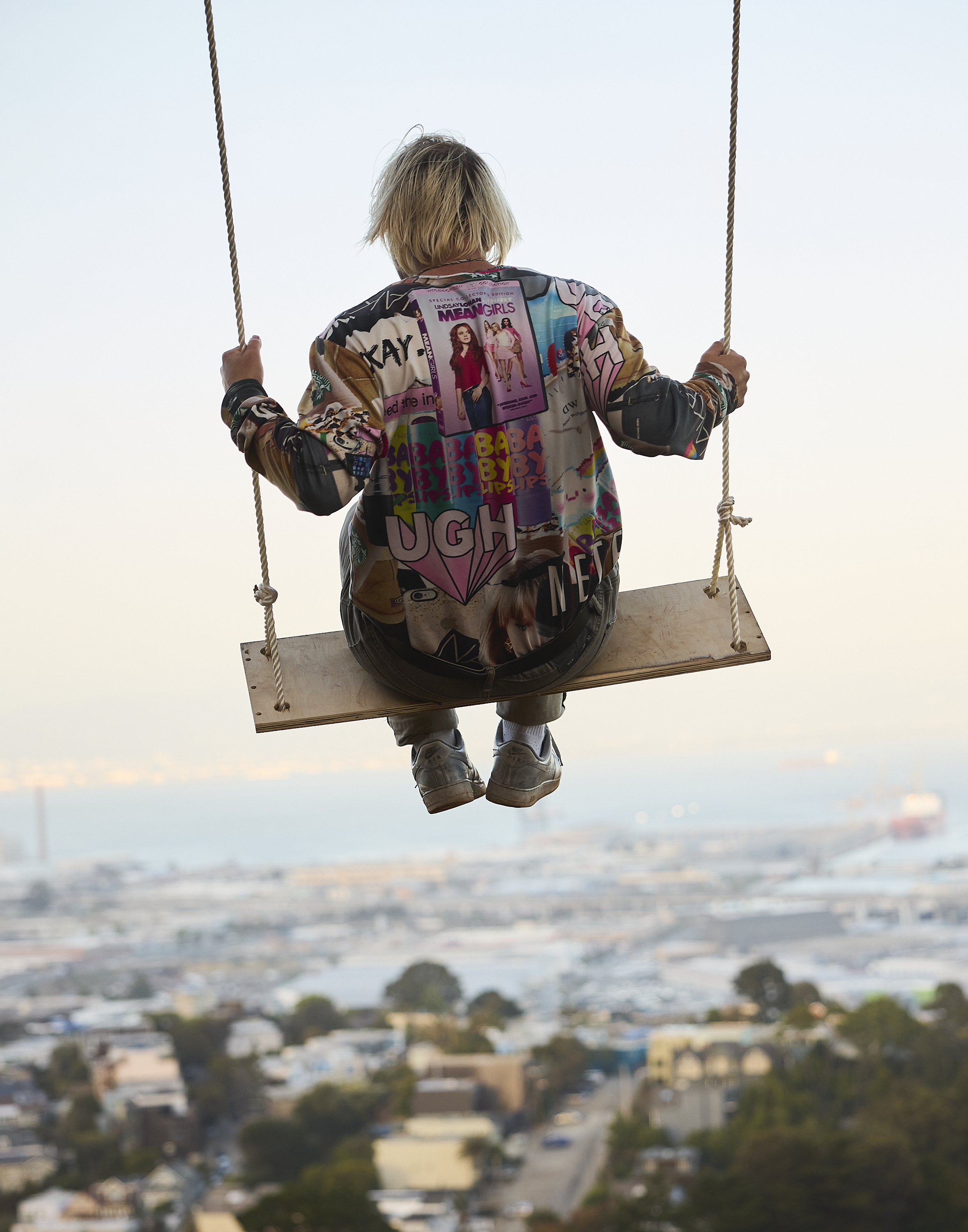 A person with blonde hair sits on a swing, facing away, overlooking a cityscape and sea. They wear a colorful jacket with patches and designs.