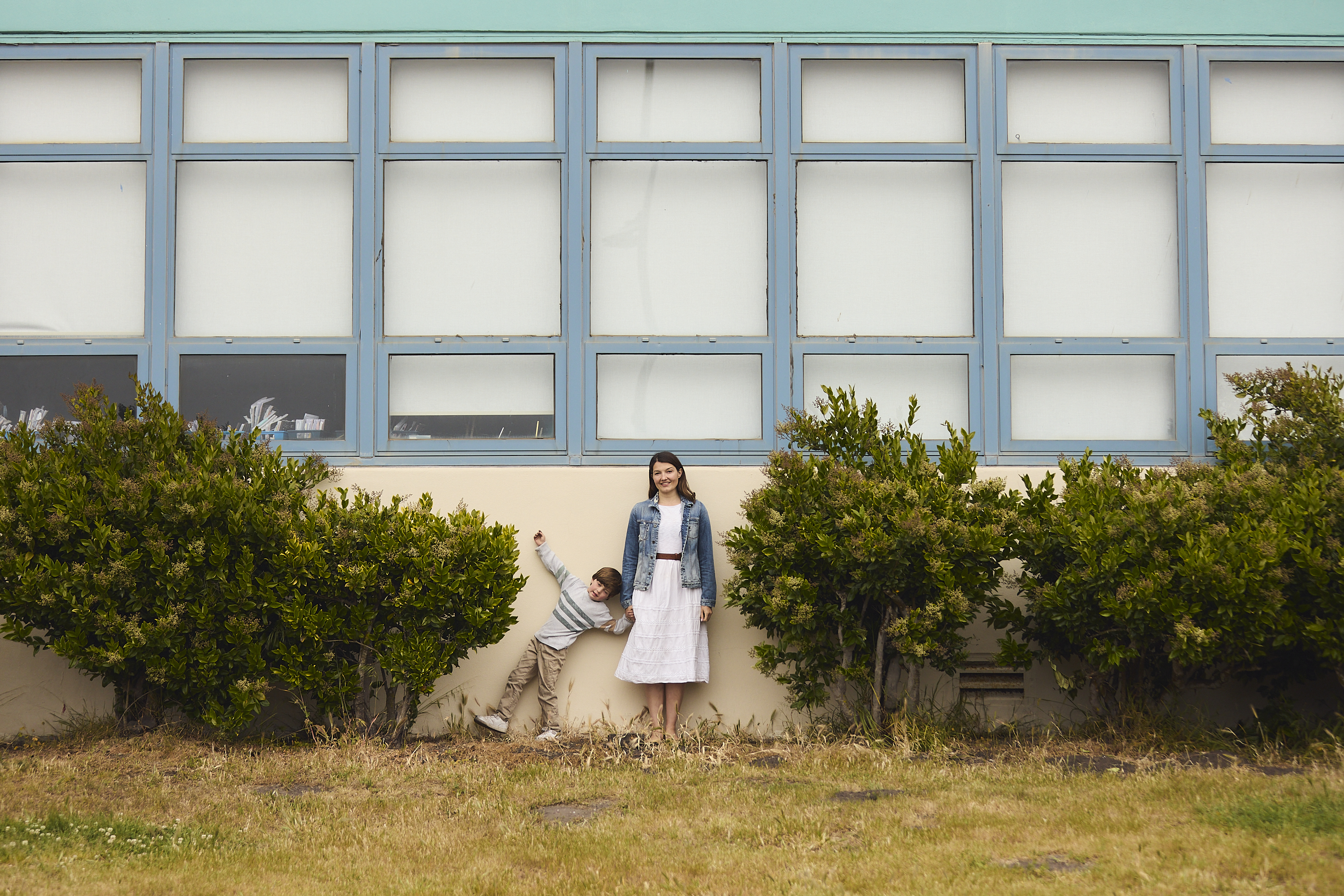 A woman and a child stand against a building with large windows, surrounded by bushes. The child leans playfully on the woman, both smiling outdoors.