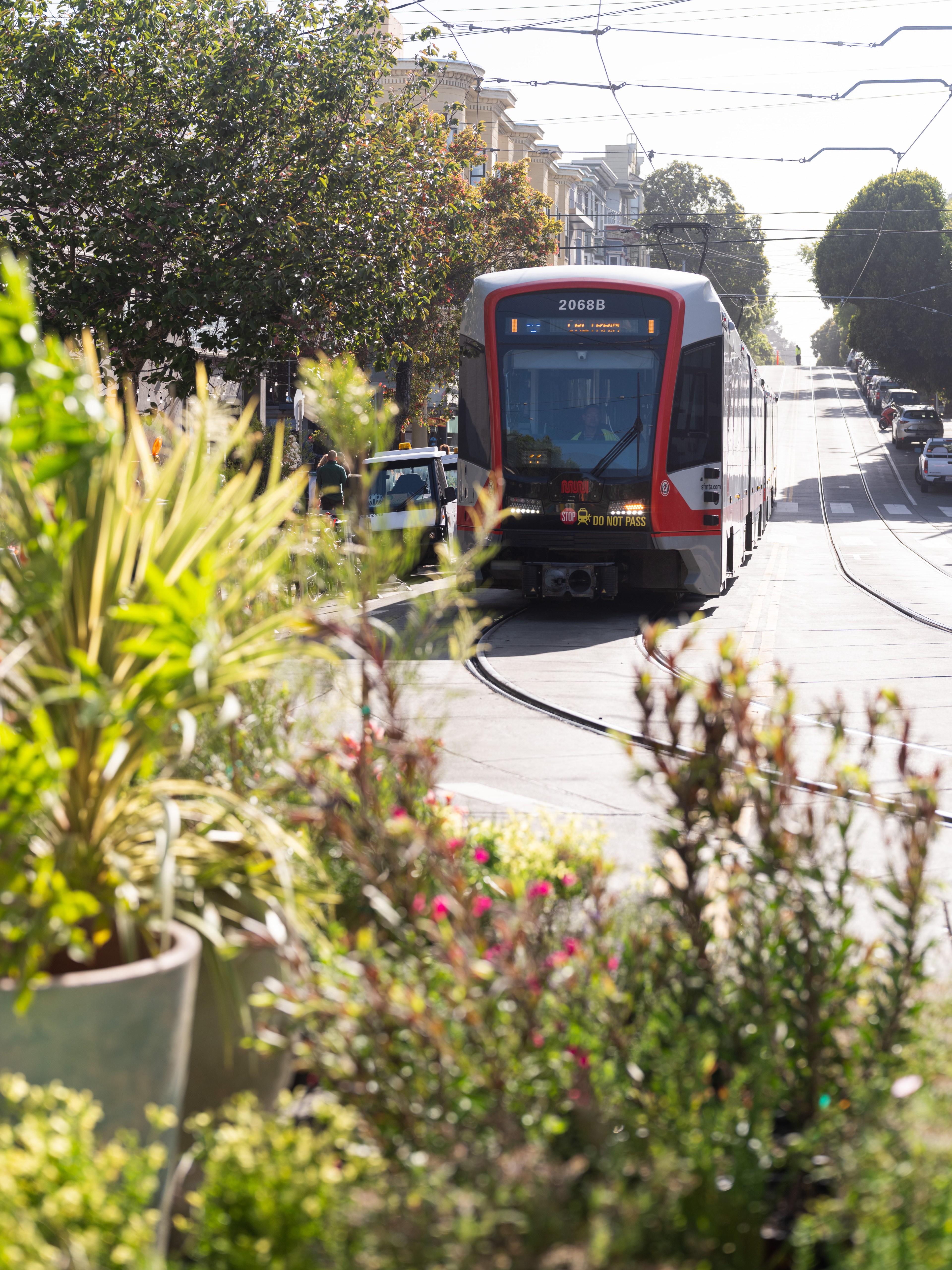 A red and white tram is moving along a city street, surrounded by trees and greenery in the foreground. The sun is shining, and buildings line the street.