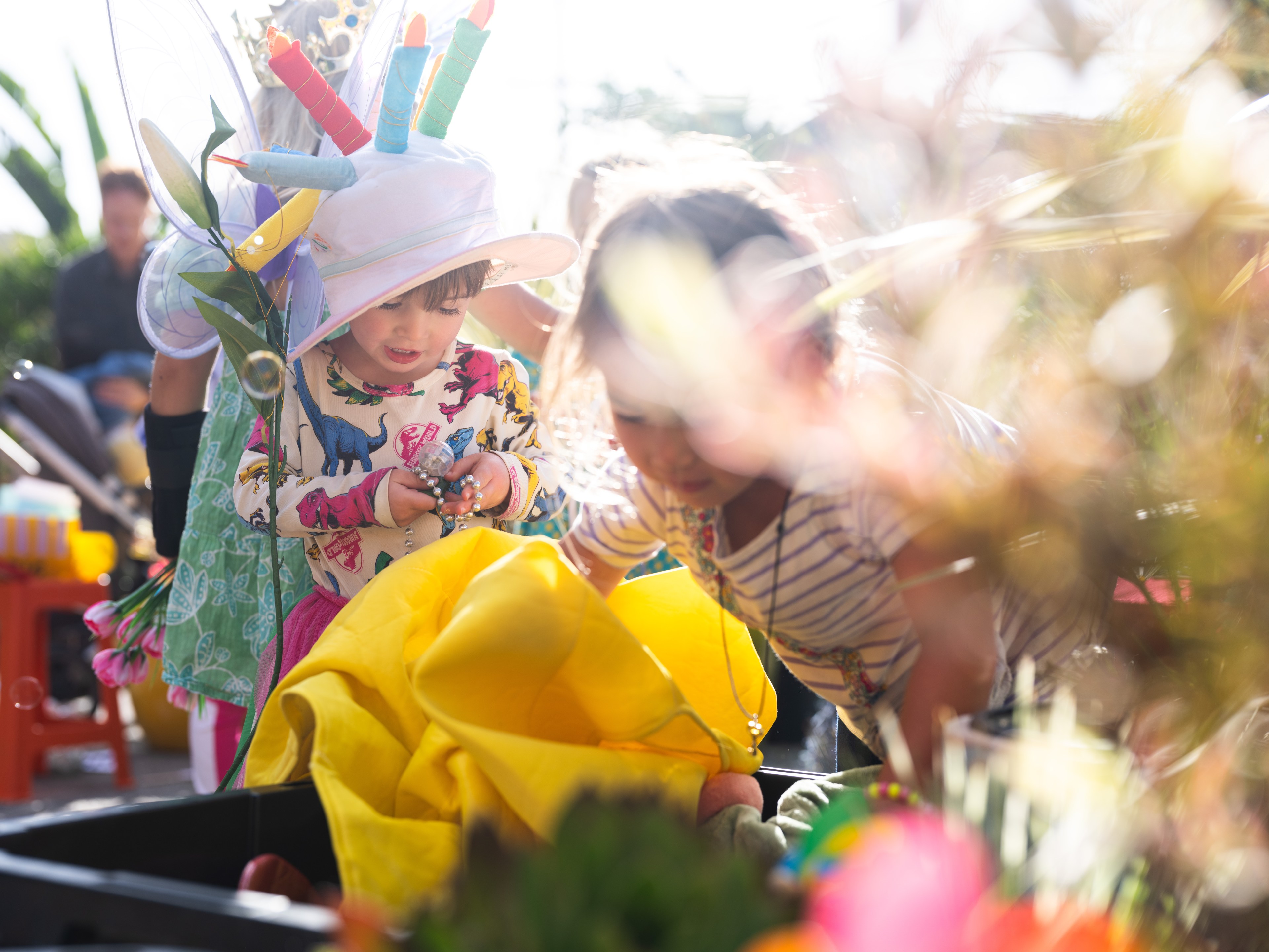 Two children are engaged in play. One wears a dinosaur-patterned shirt and a colorful hat, surrounded by vibrant objects and blurred foliage in sunlight.