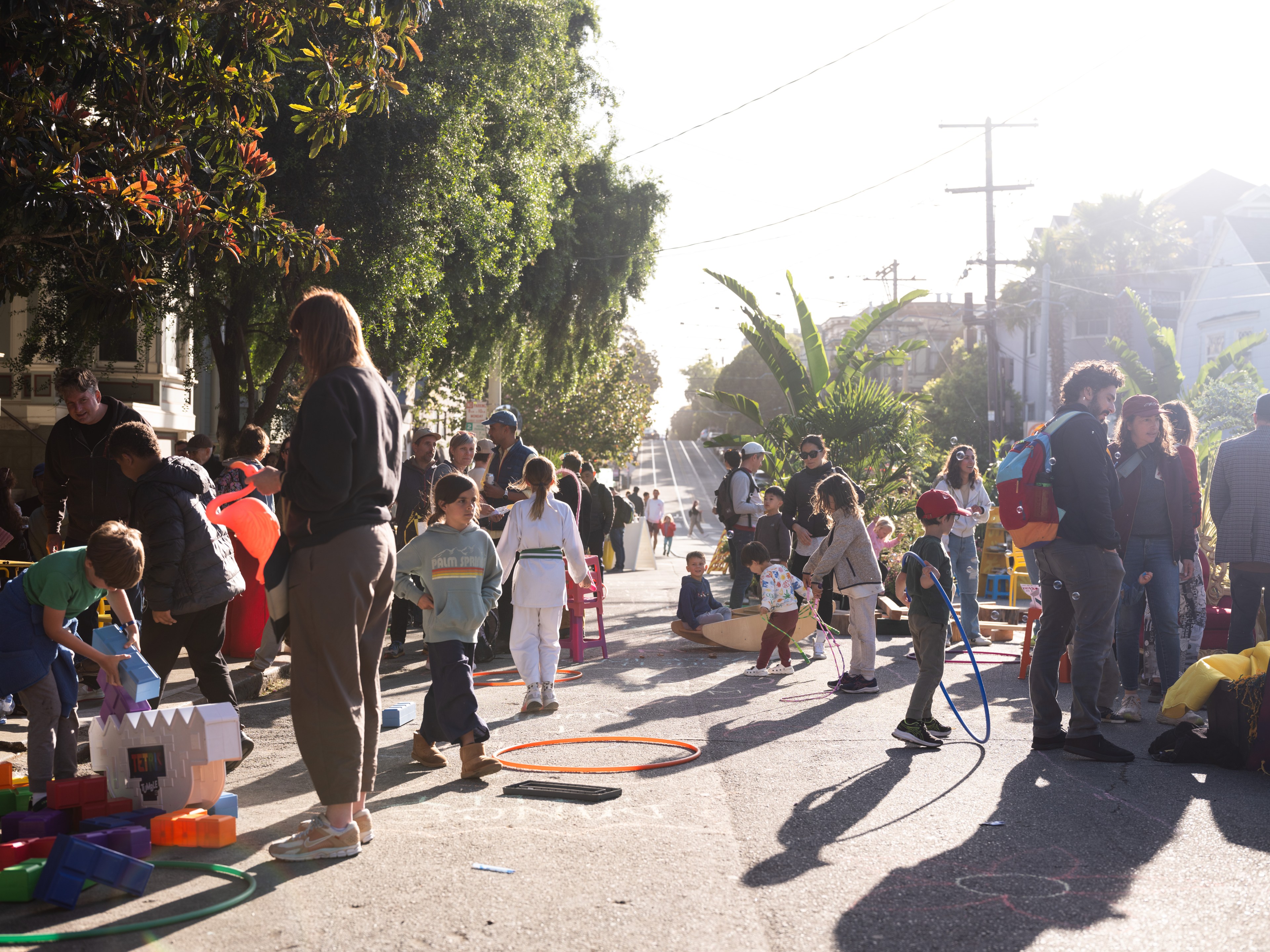 The image shows a lively street scene with children and adults playing and interacting, surrounded by trees and buildings. Toys and brightly colored hoops are scattered around.