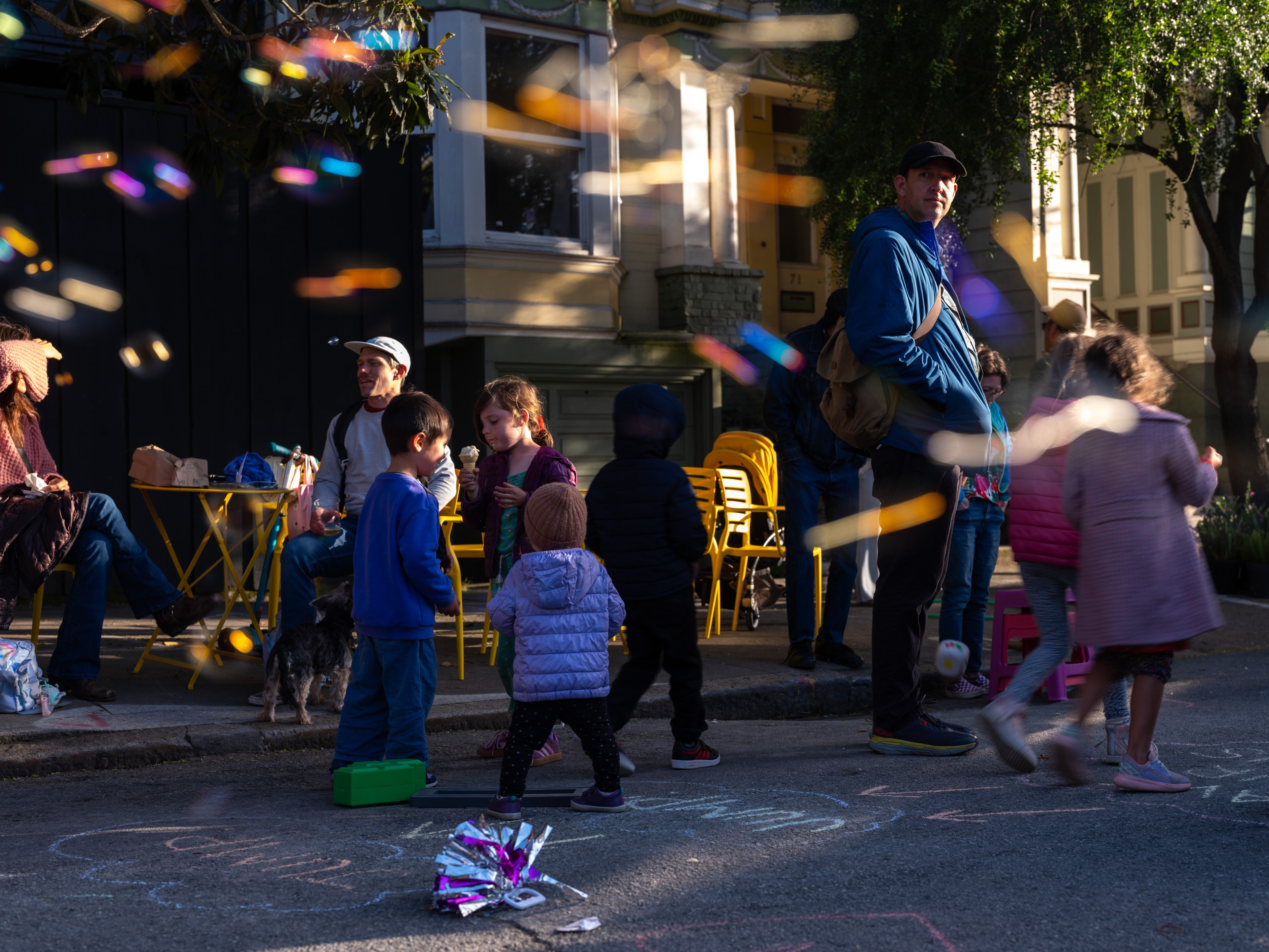 A lively street scene shows children playing with chalk, surrounded by colorful blurred lights. Adults sit at yellow tables. A dog and vibrant chairs are visible.
