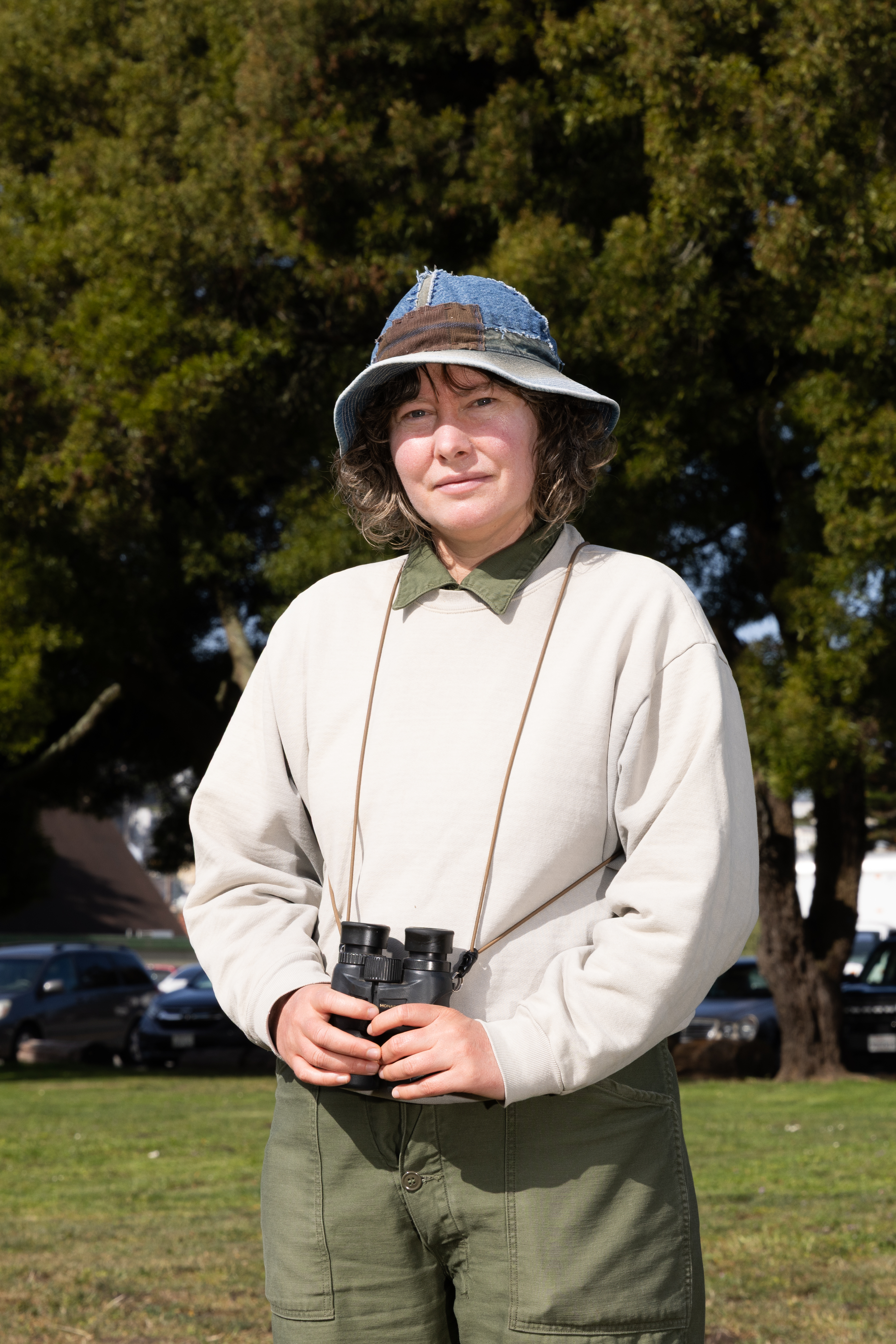A person stands outdoors, wearing a patched denim bucket hat and a light sweatshirt. They hold binoculars and are in front of trees and parked cars.
