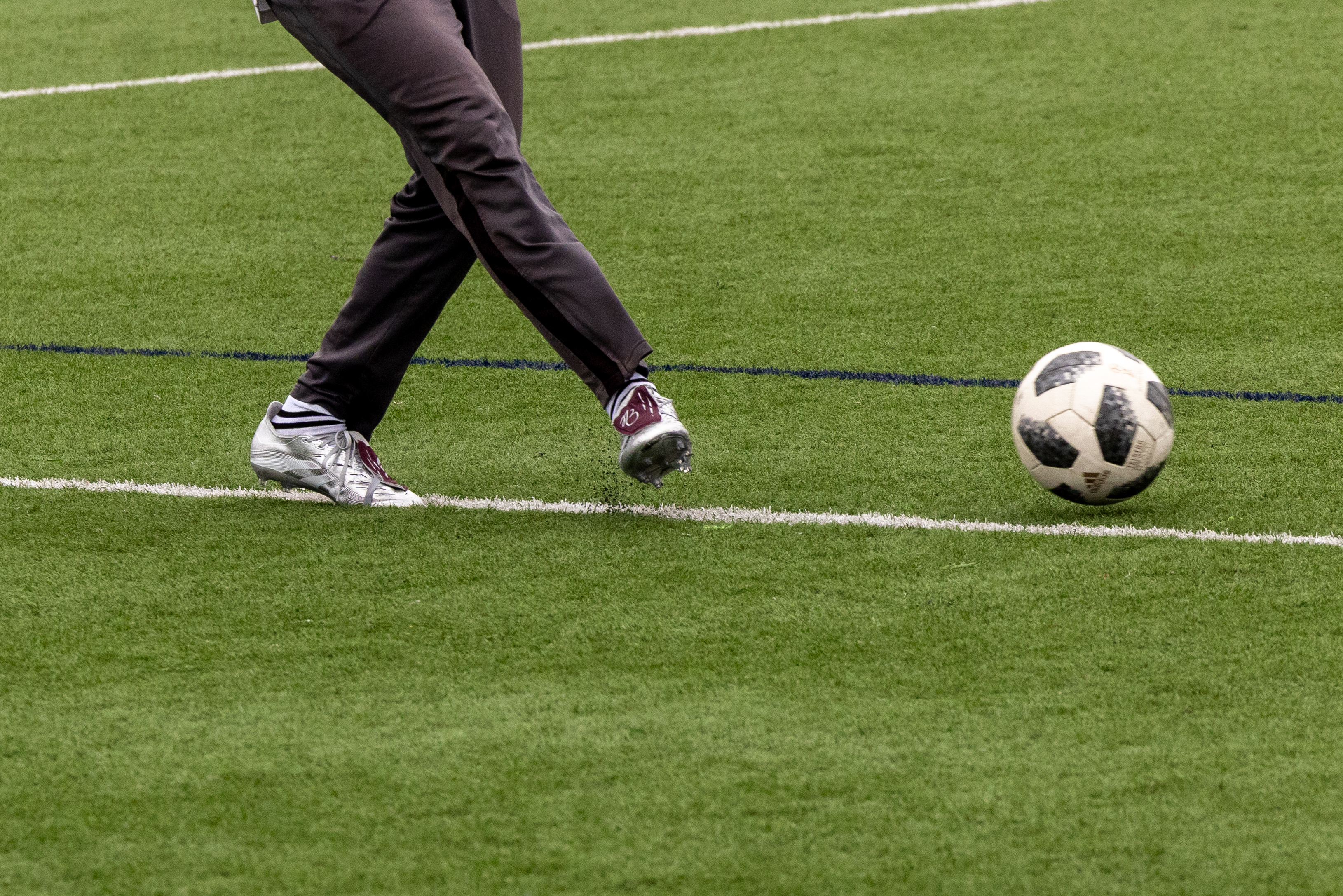 A person wearing dark pants and white shoes kicks a black and white soccer ball on a green grass field with white lines.