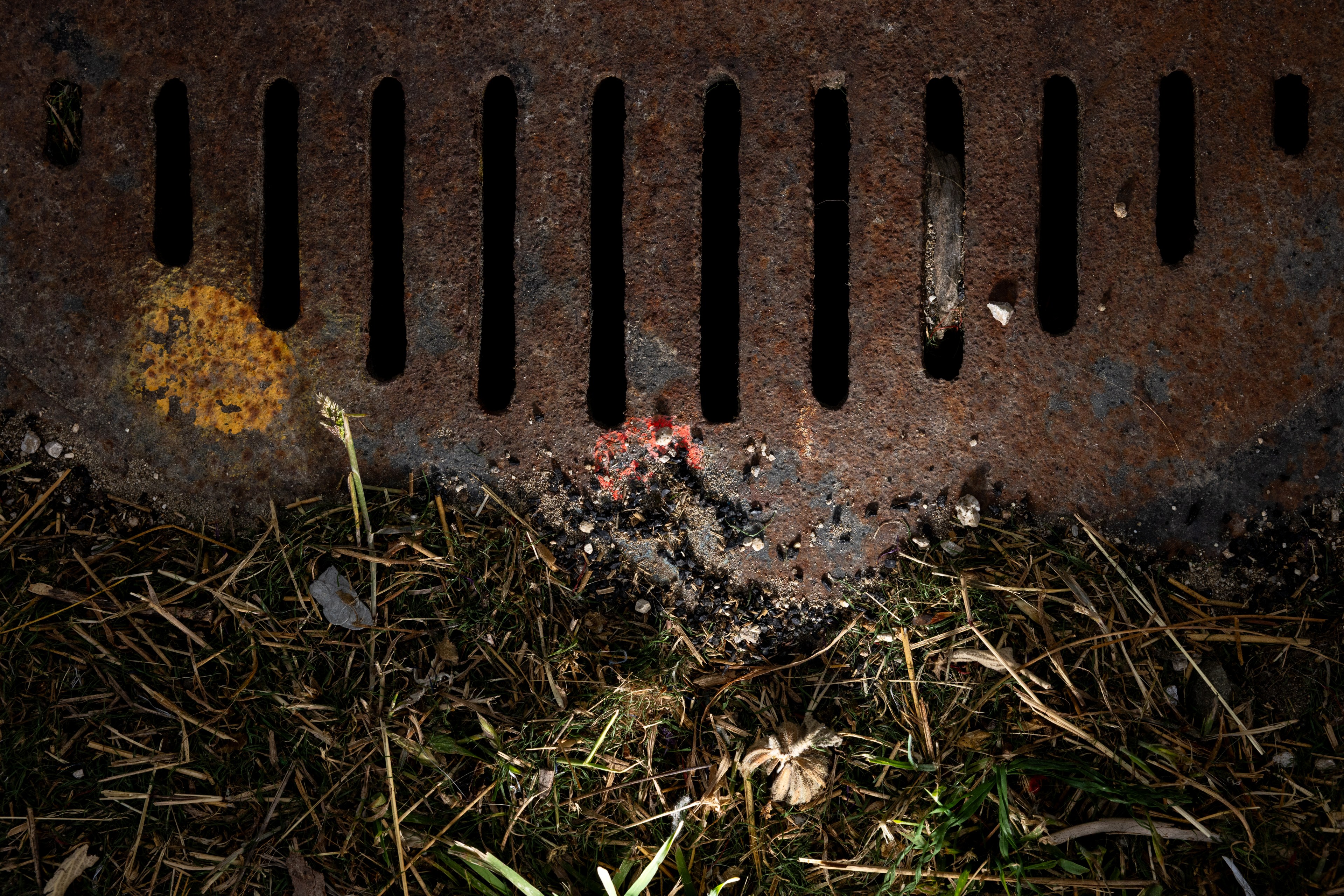 A rusty metal drain cover with vertical slits lies on grass. A yellow and a red mark are visible on the metal. Pebbles and debris surround the edge.