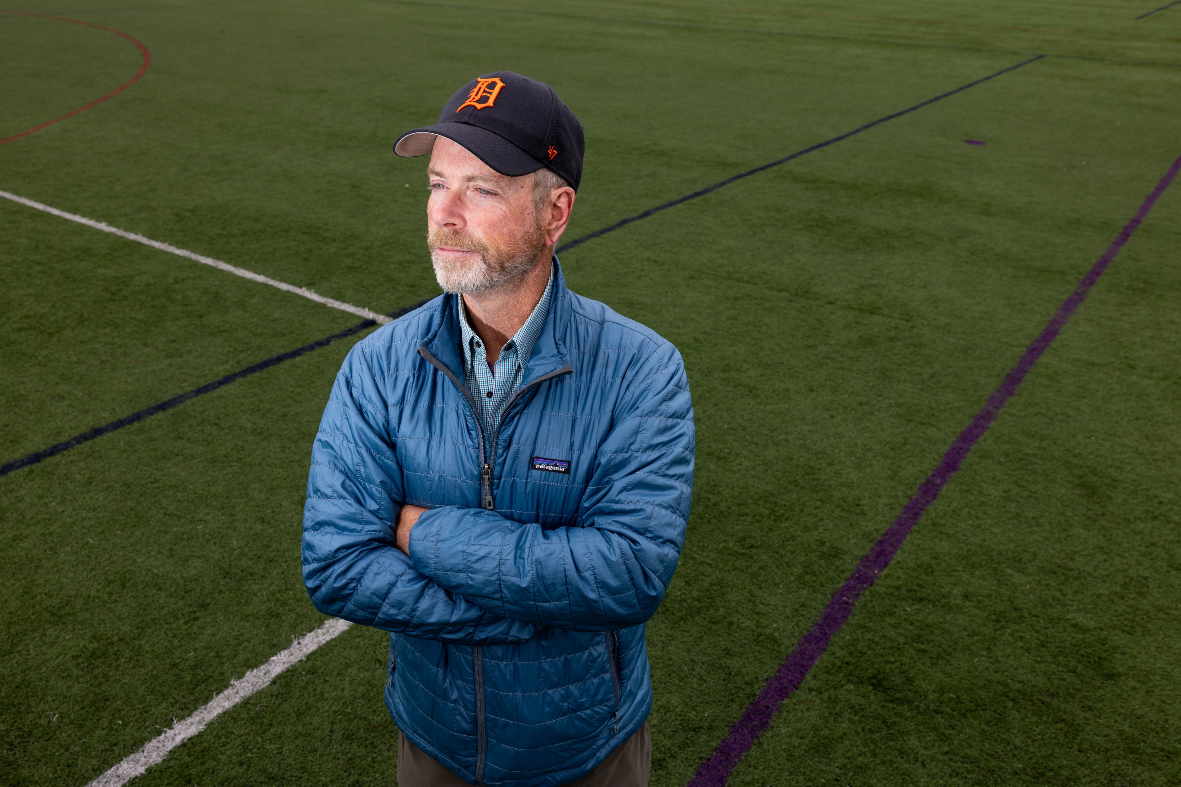 A man stands on a sports field with crossed arms, wearing a blue jacket and a black cap. The field has various colored lines on the green grass.