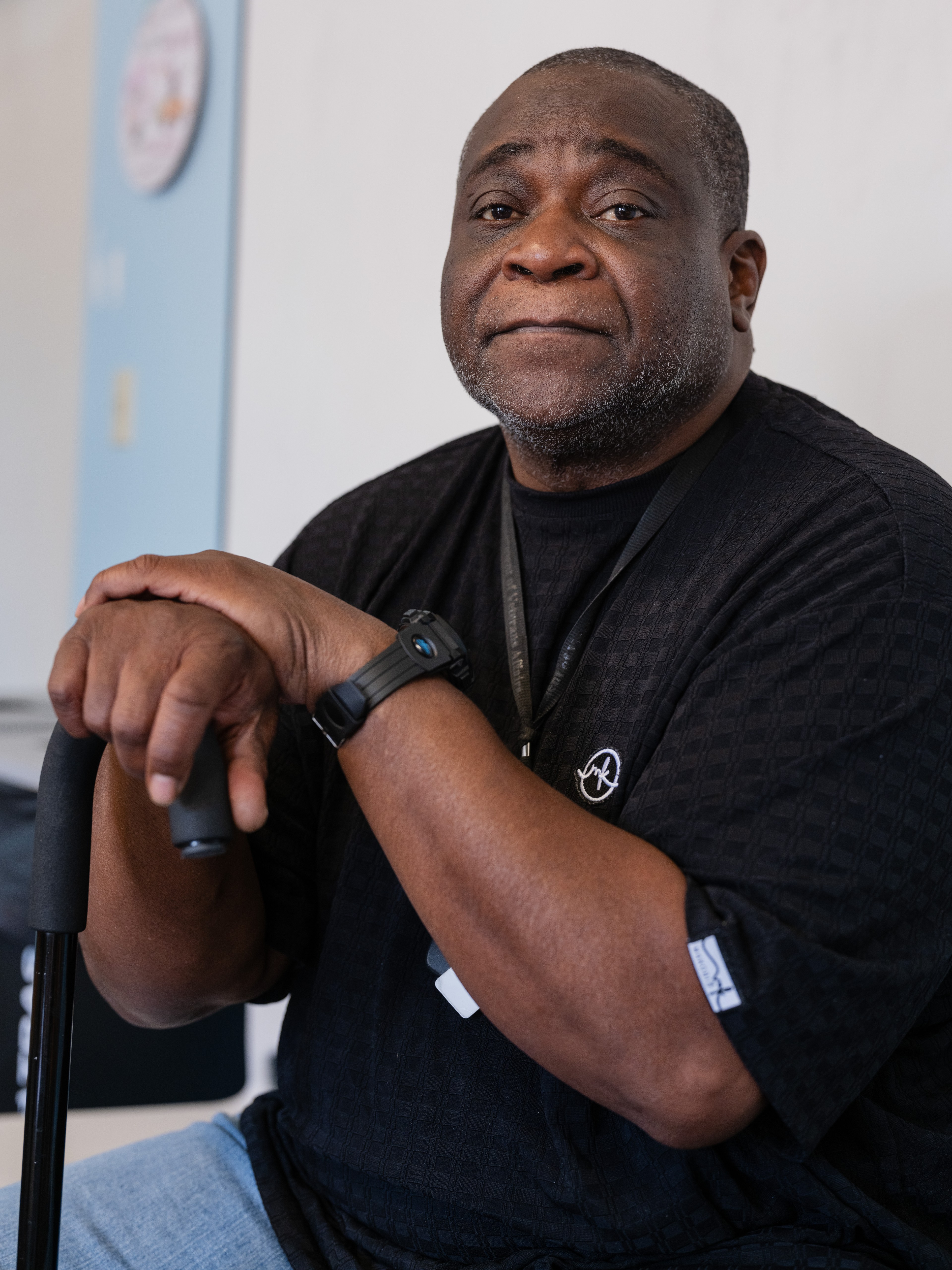 A man sits, resting on a cane, wearing a black shirt and watch. The background shows a light blue and white wall with a round object on it.