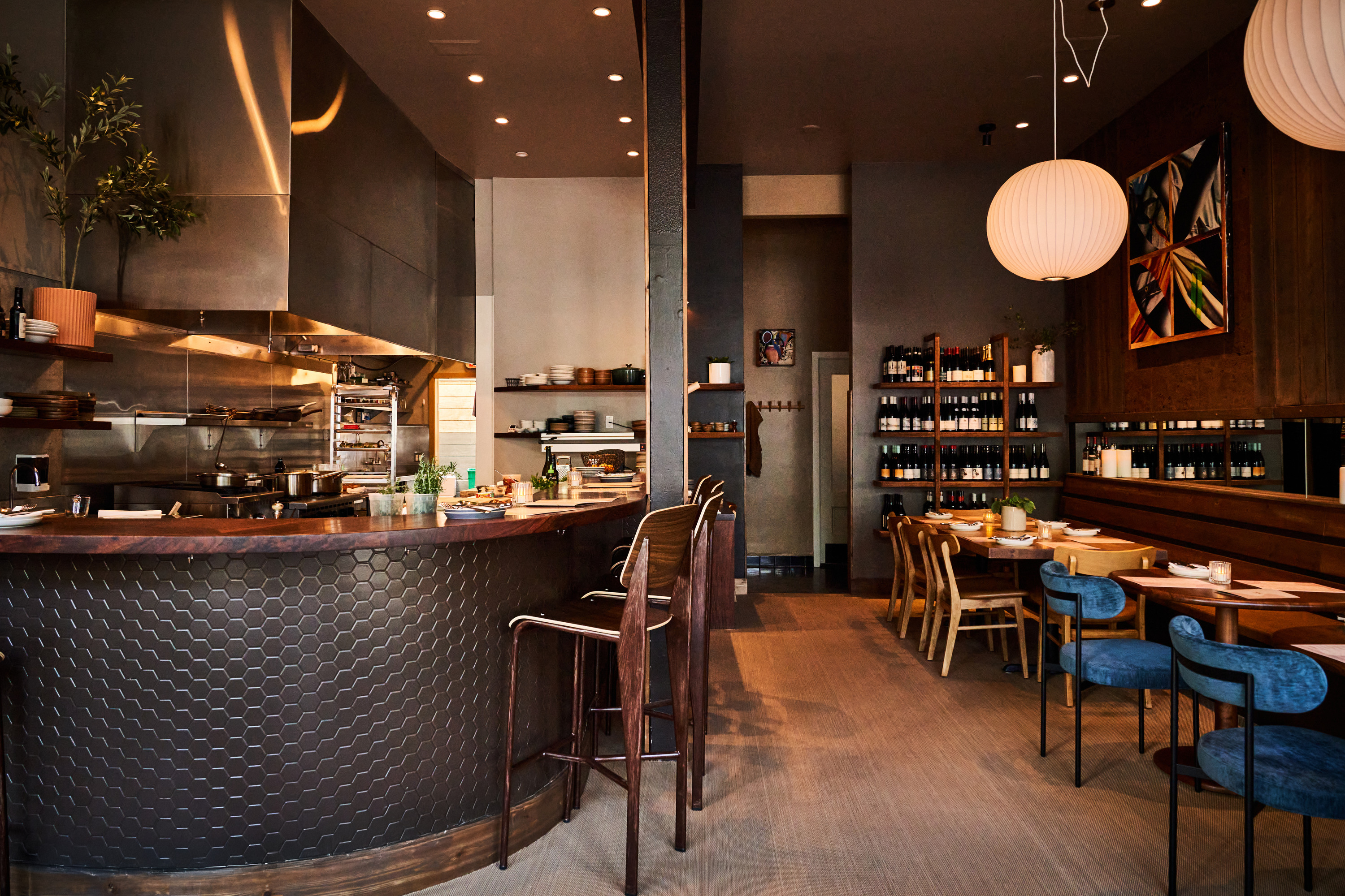 The image shows a cozy restaurant interior with a hexagonal-tiled bar, wooden stools, round pendant lights, shelves of wine, and neatly set tables.