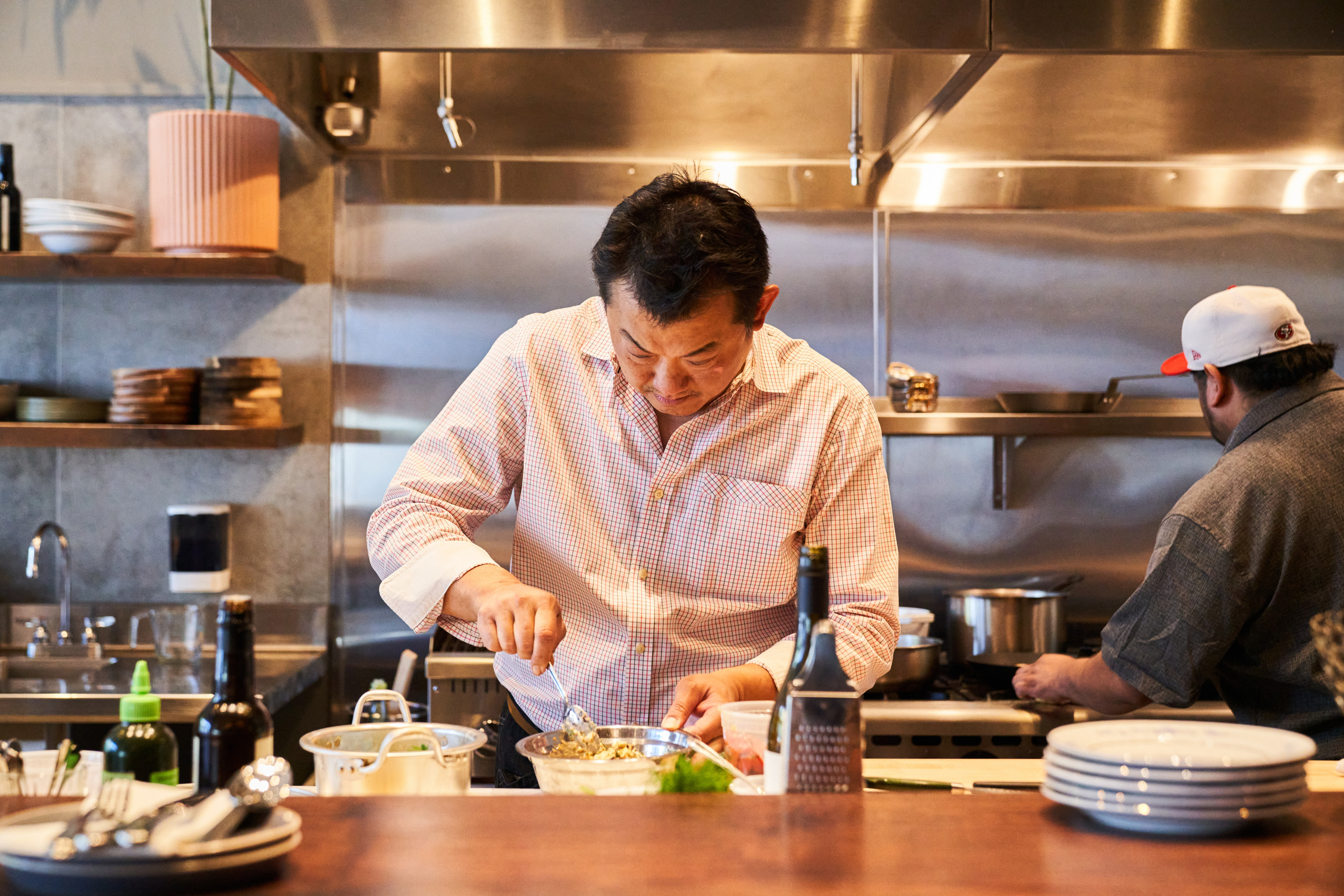 Two chefs are in a professional kitchen. One is focusing on mixing ingredients in a bowl, while the other is cooking at the stove. Shelves with dishes are visible.